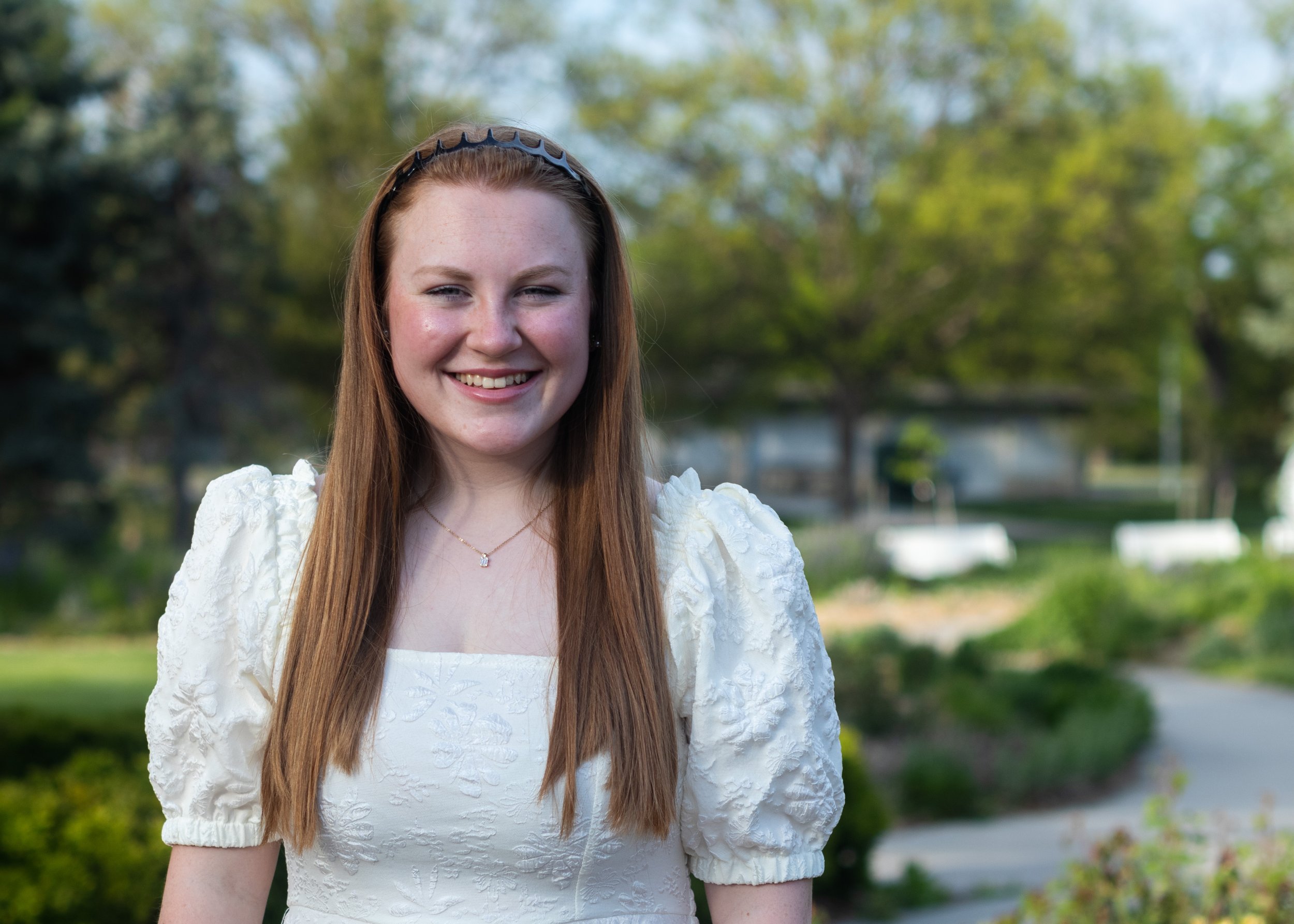 A young woman with long reddish-brown hair, smiling outdoors, wearing a white textured dress and a black headband, with a background of green trees and a park.