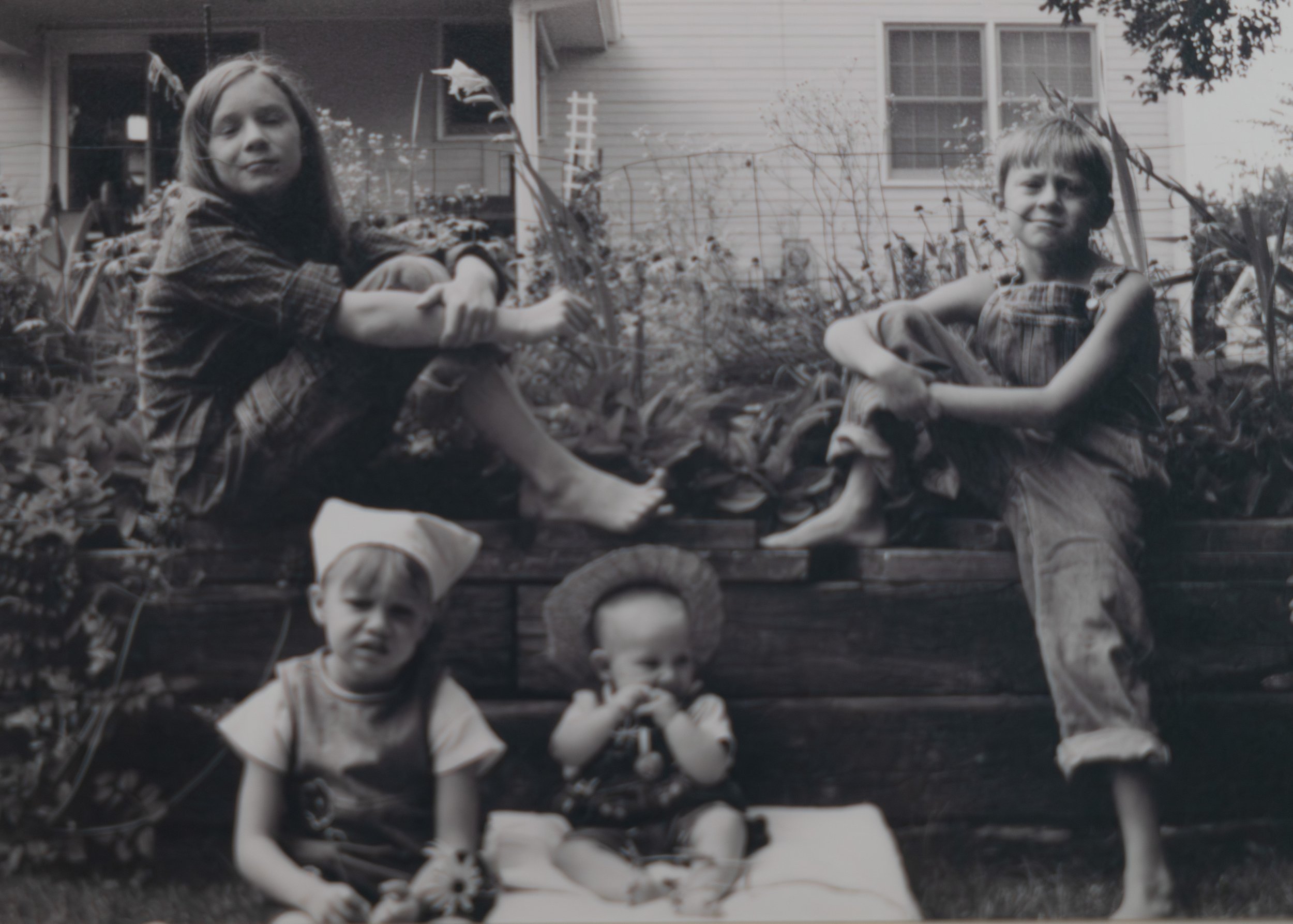 Black-and-white photo of five children sitting outdoors in a garden, with a house in the background, some with serious or neutral expressions, wearing farm style clothing, and plants around them.