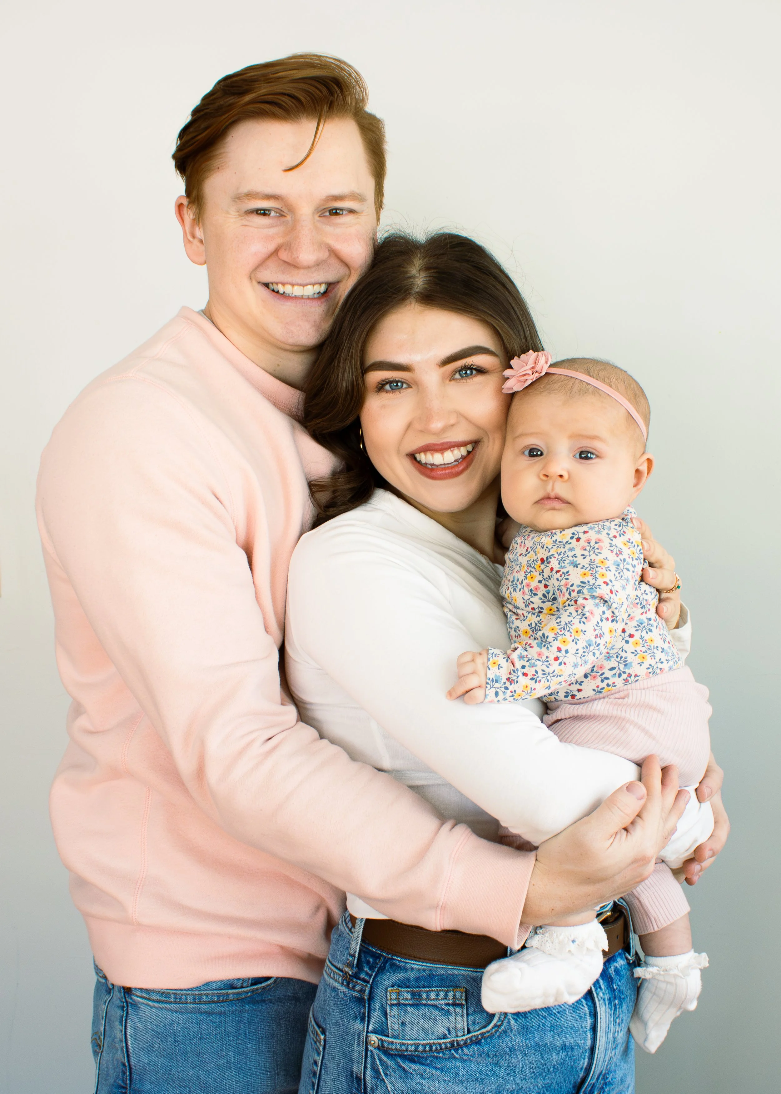 A happy family of three with a man, woman, and baby girl posing together against a neutral background.