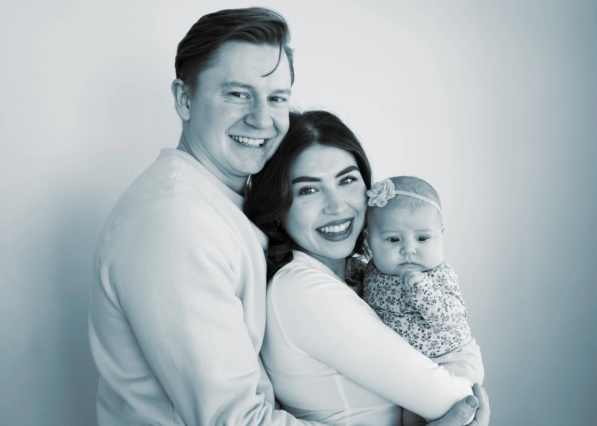 A happy family portrait of a man, woman, and baby, all smiling and close together against a plain background.