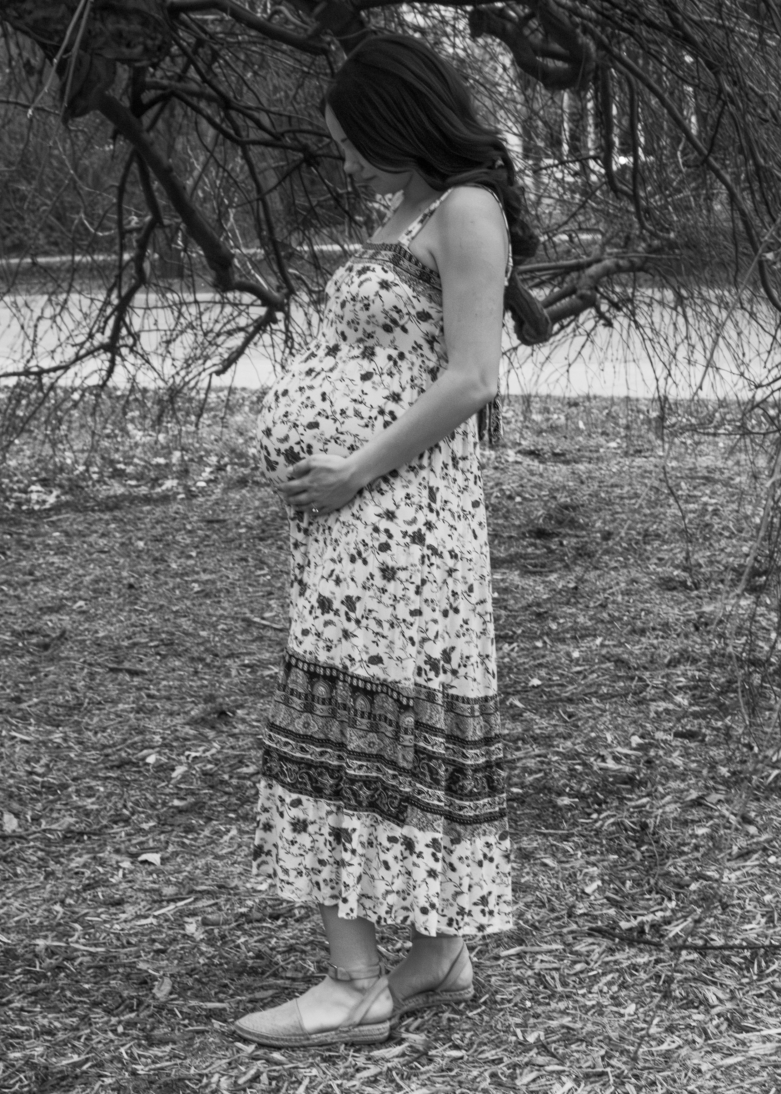 A woman with long dark hair standing outdoors under tree branches, touching her pregnant belly, dressed in a floral dress and sandals, in a black-and-white photo.