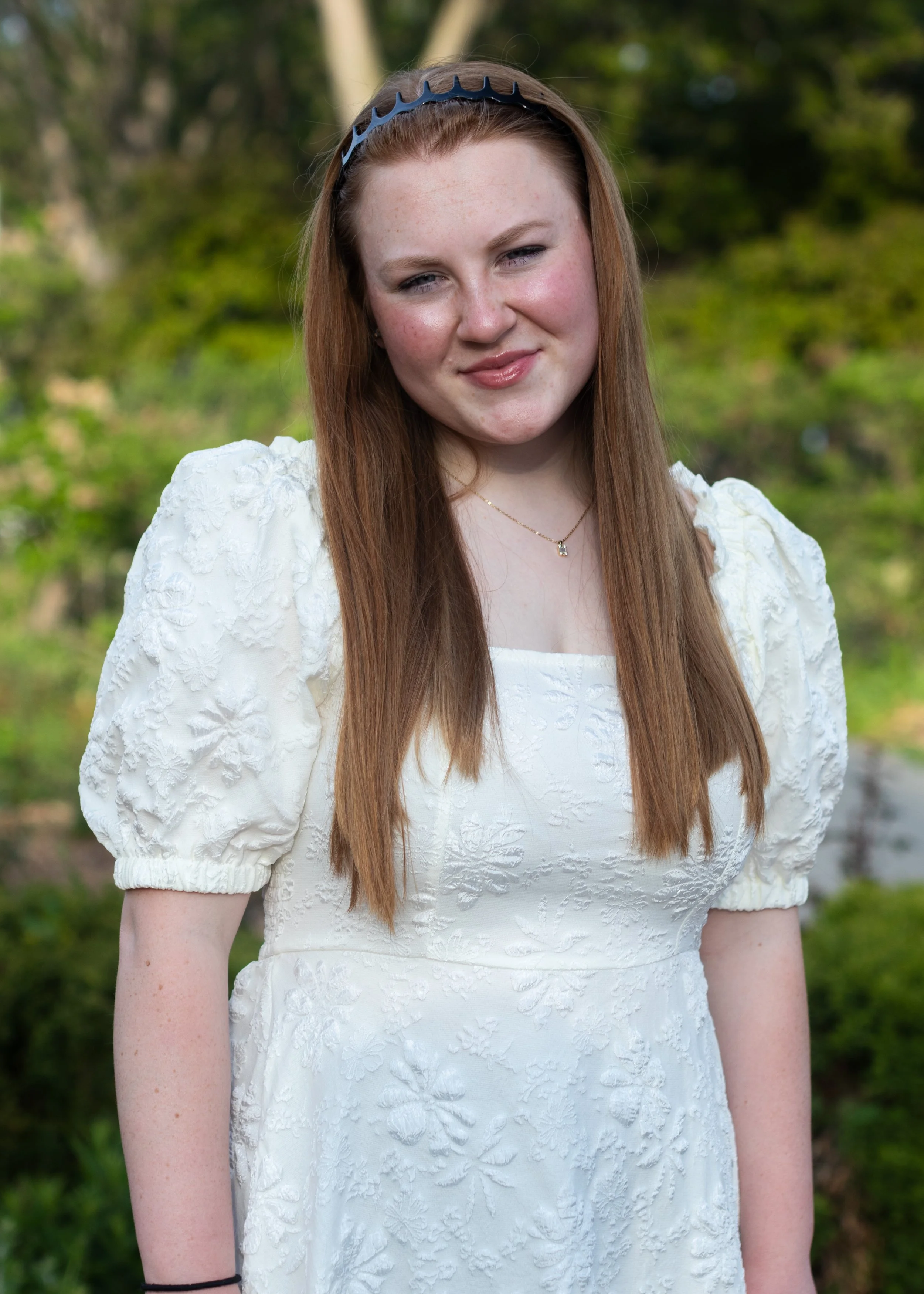 A young woman with long red hair wearing a white embroidered dress and a black headband, standing outdoors with a blurred green background.