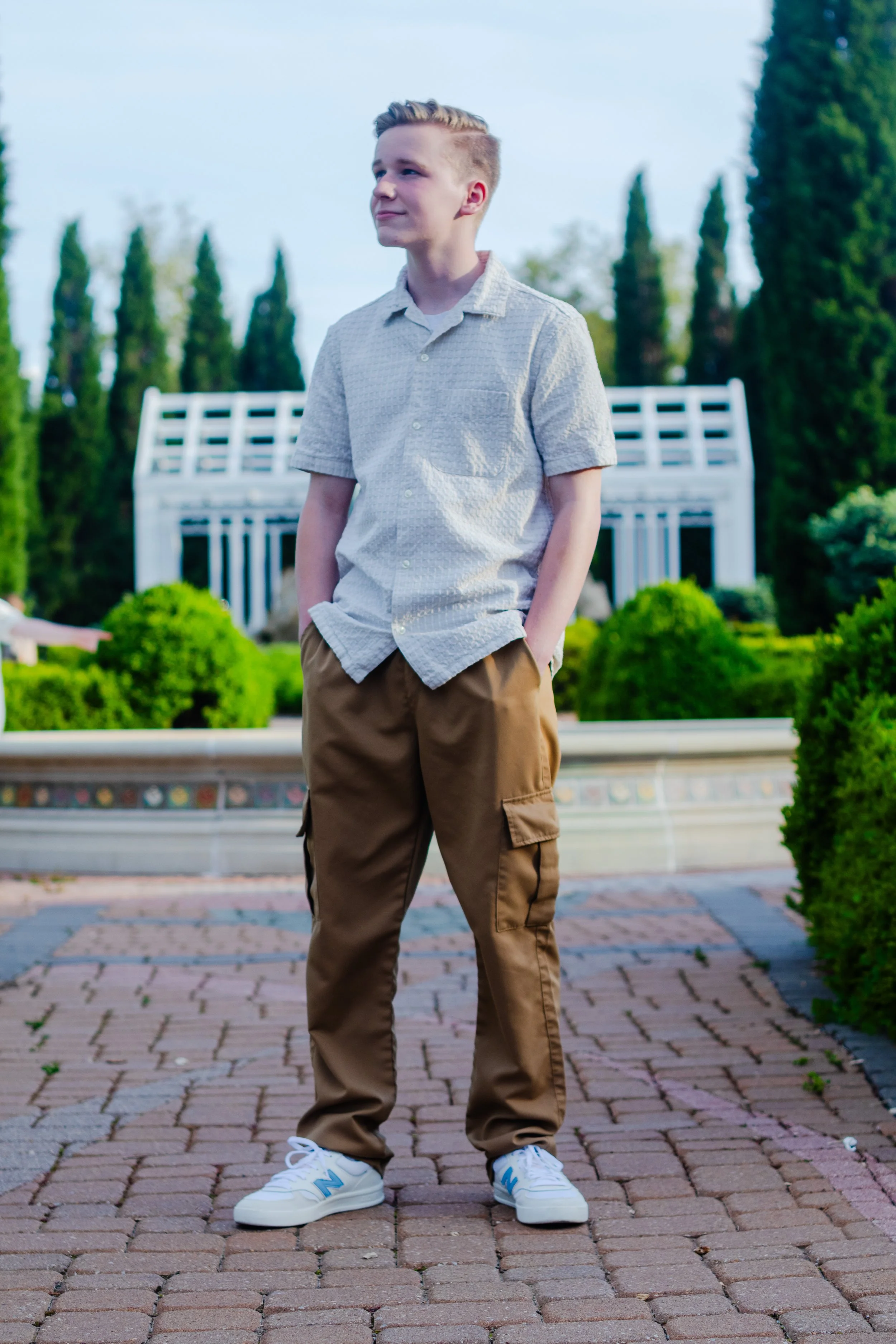 A teenage boy with short blonde hair, wearing a light gray short-sleeved button-up shirt, brown pants, and white sneakers, standing outdoors on a brick pathway with greenery and tall trees in the background.