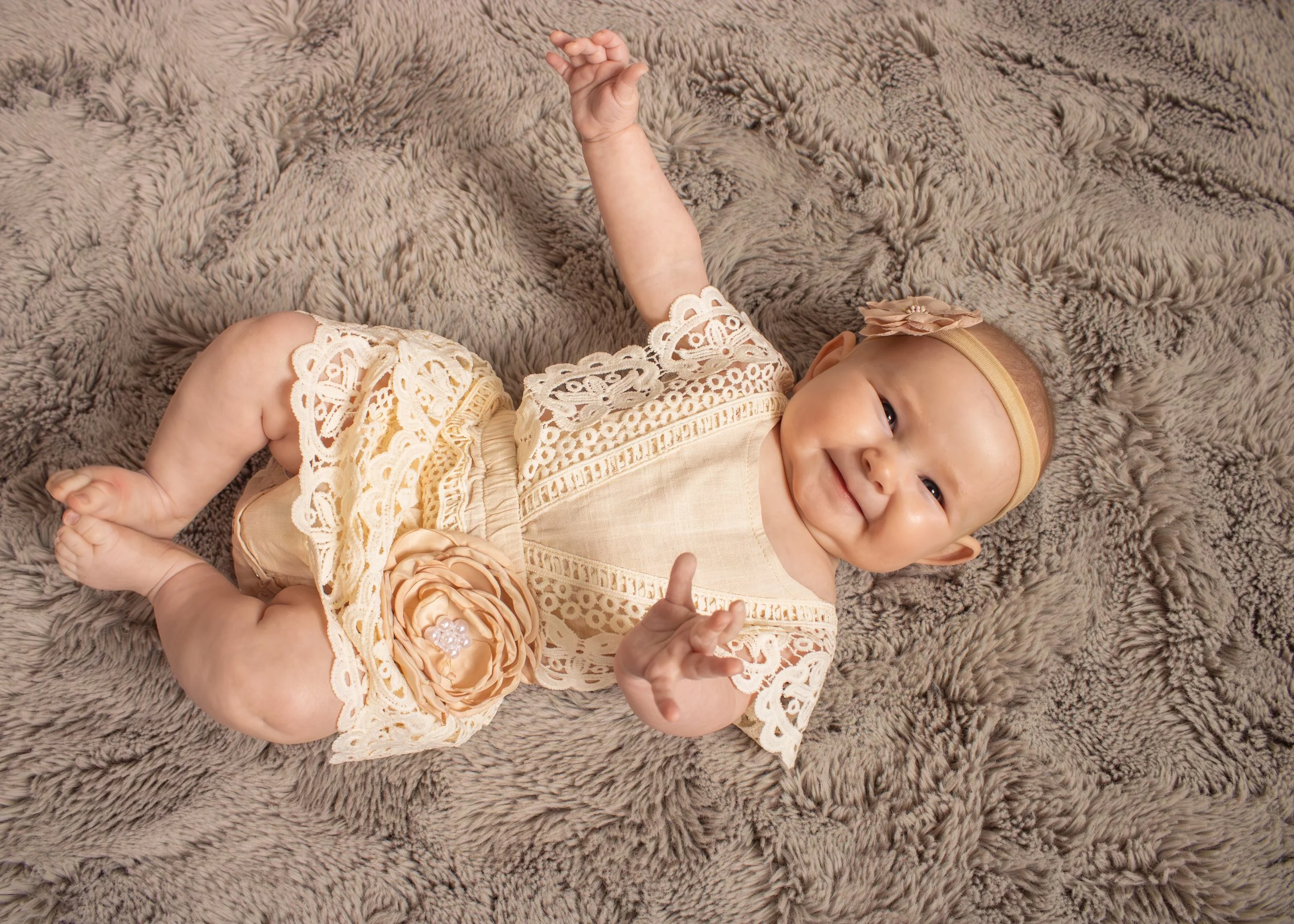 A smiling baby girl lying on a plush, textured gray blanket, wearing a cream-colored lace dress with peach accents and a peach headband with a bow.