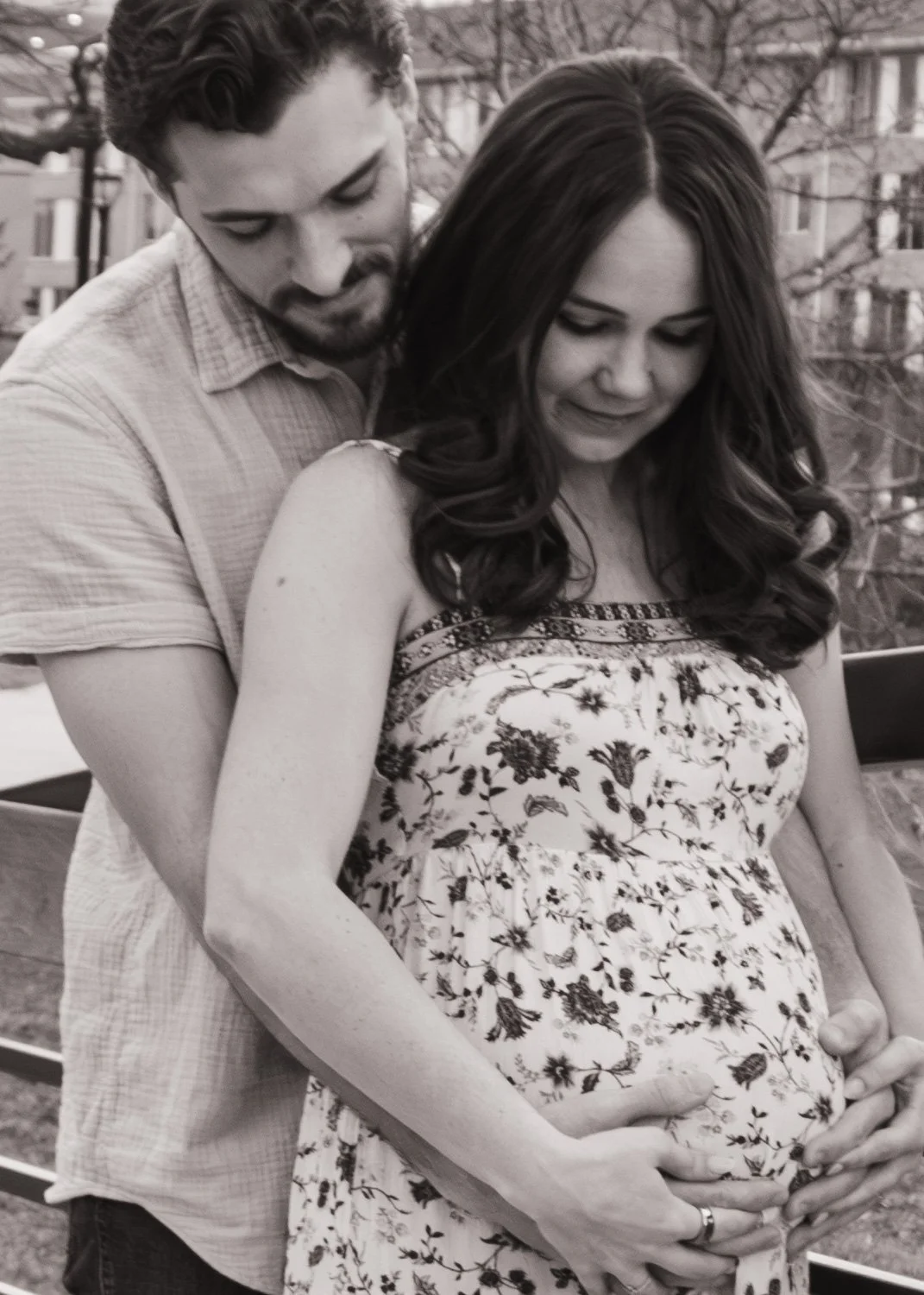 A pregnant woman with long dark hair wearing a floral dress, is cradling her baby bump, with a man standing behind her, both smiling softly and looking down at her belly. The background shows trees and apartment buildings.
