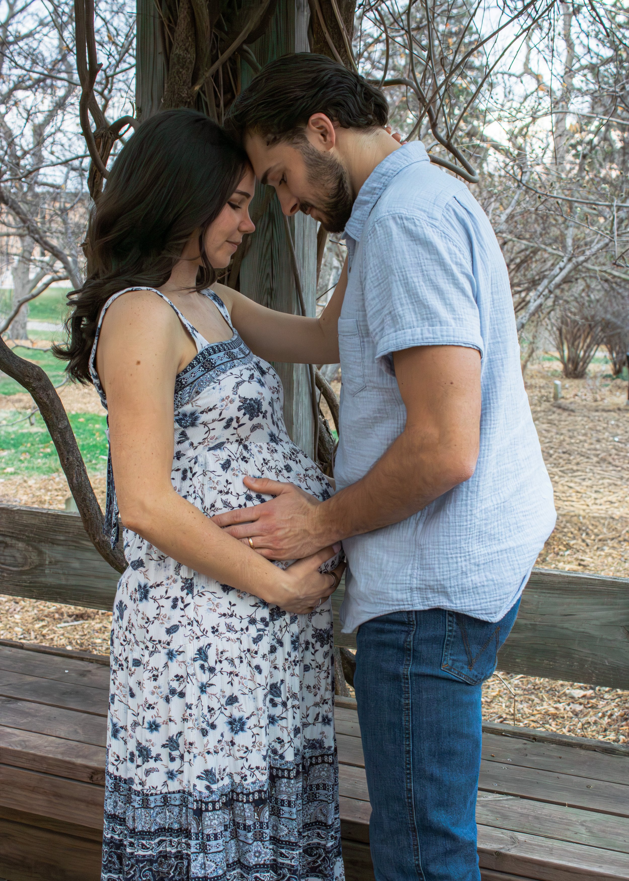 A pregnant woman and a man touching foreheads with their eyes closed, standing close outdoors next to a tree with bare branches, during daytime.