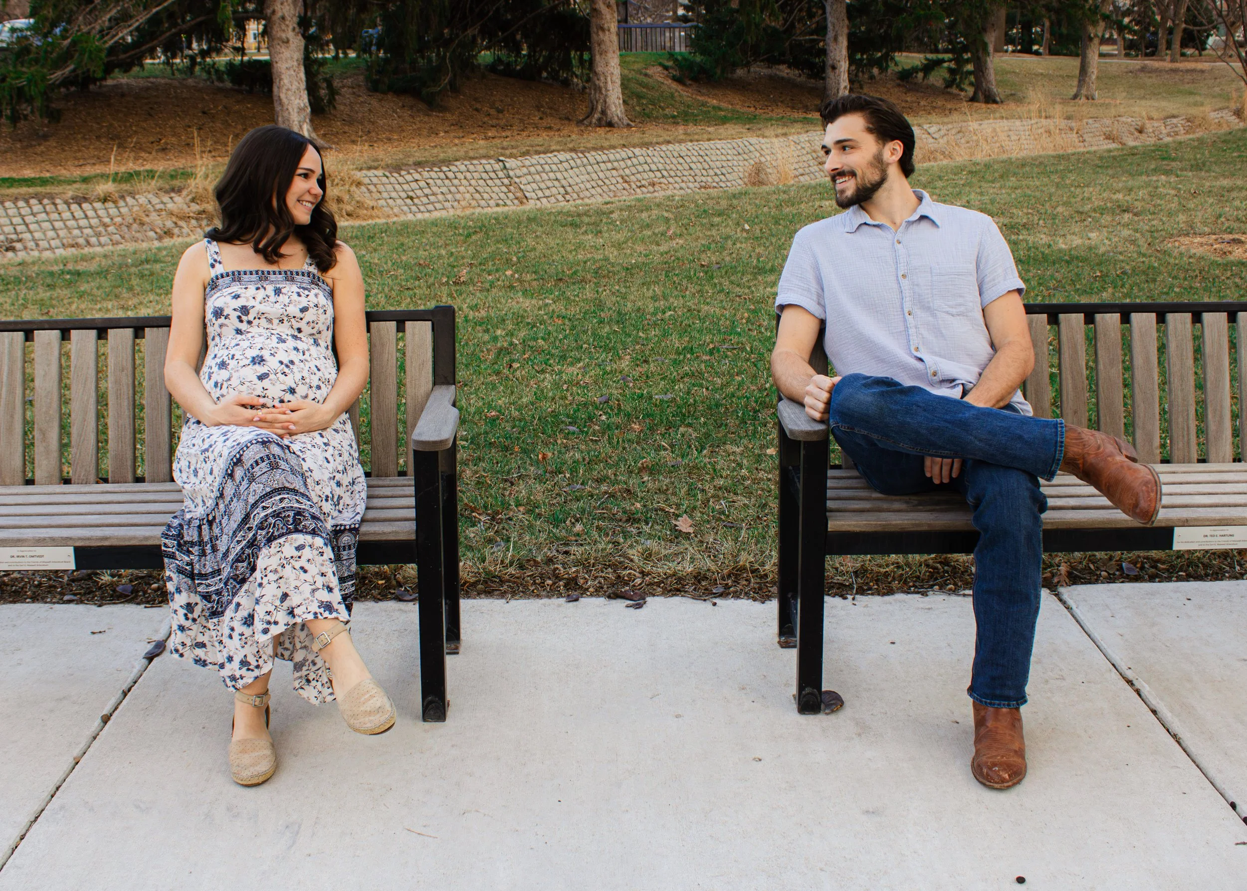A man and a woman sitting on separate benches, smiling and looking at each other in a park with trees and grass.