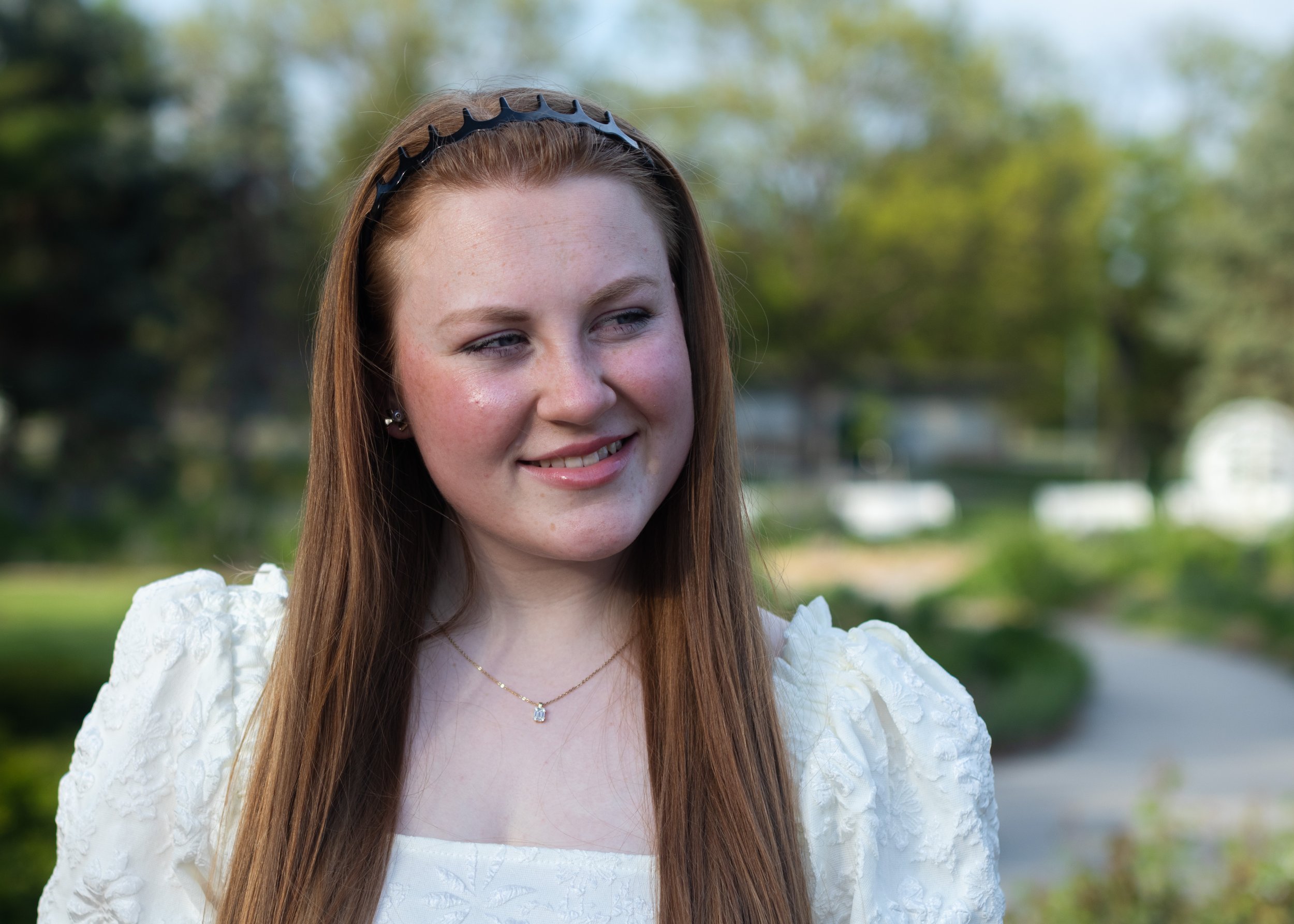 Close-up of a young woman with long red hair, wearing a white dress with puffed sleeves, a black headband, and jewelry, standing outdoors with blurred green trees in the background.
