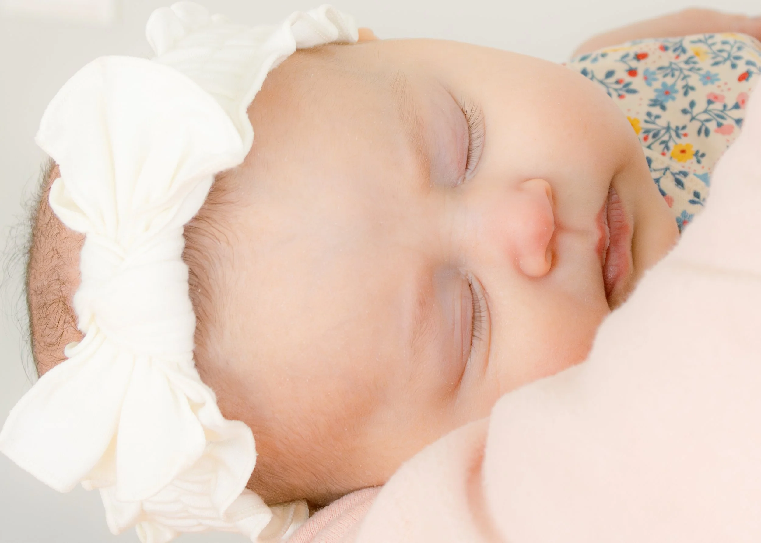 Close-up of a sleeping baby with fair skin, wearing a floral-patterned outfit, lying on a white surface, with a swaddle or blanket partially covering their head.