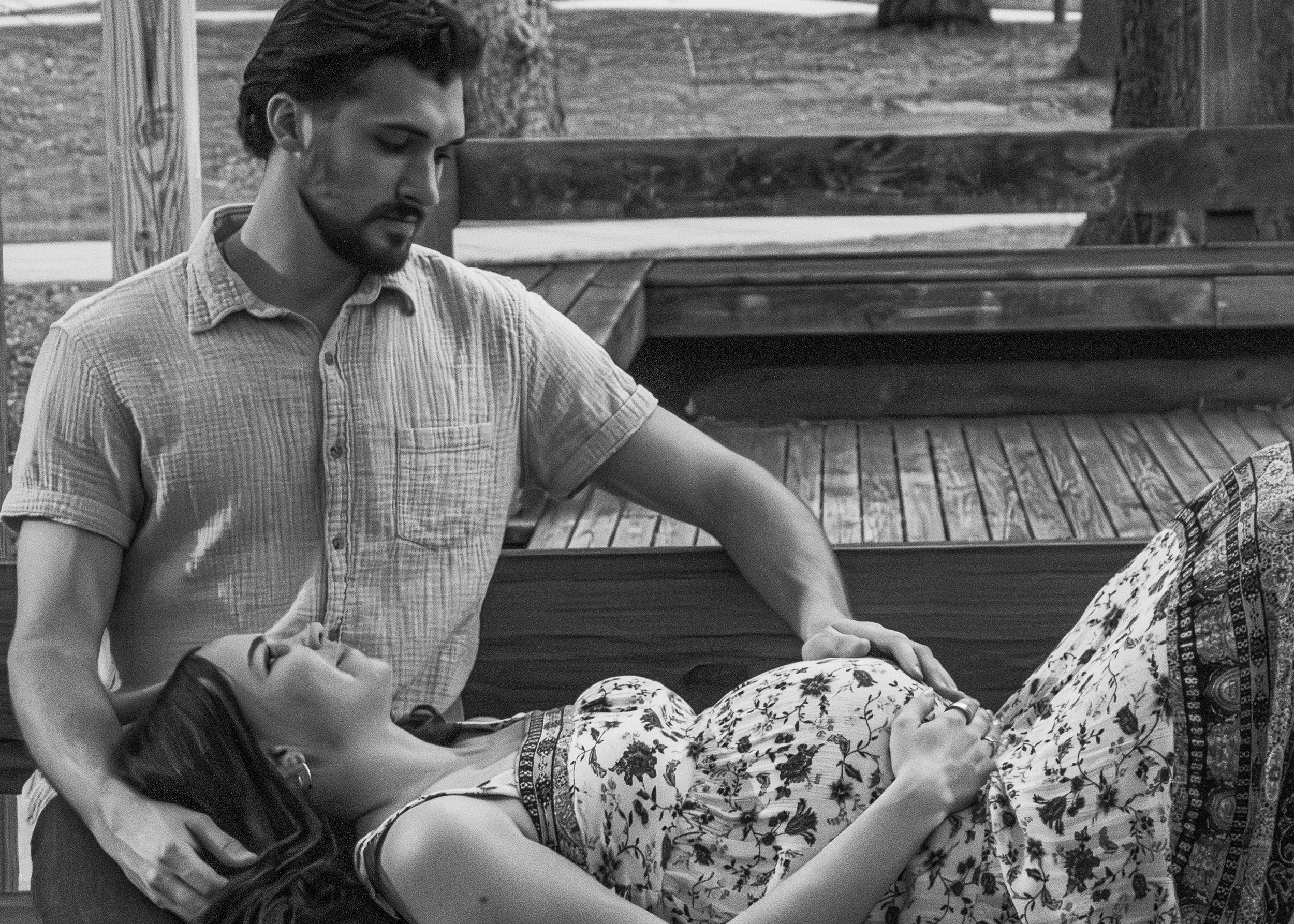 A young man with dark hair and a beard, wearing a plaid shirt, is sitting on a park bench, looking down at something on his lap, with a woman in a floral dress sitting beside him. The scene is outdoors with trees and wooden park benches in the backgr