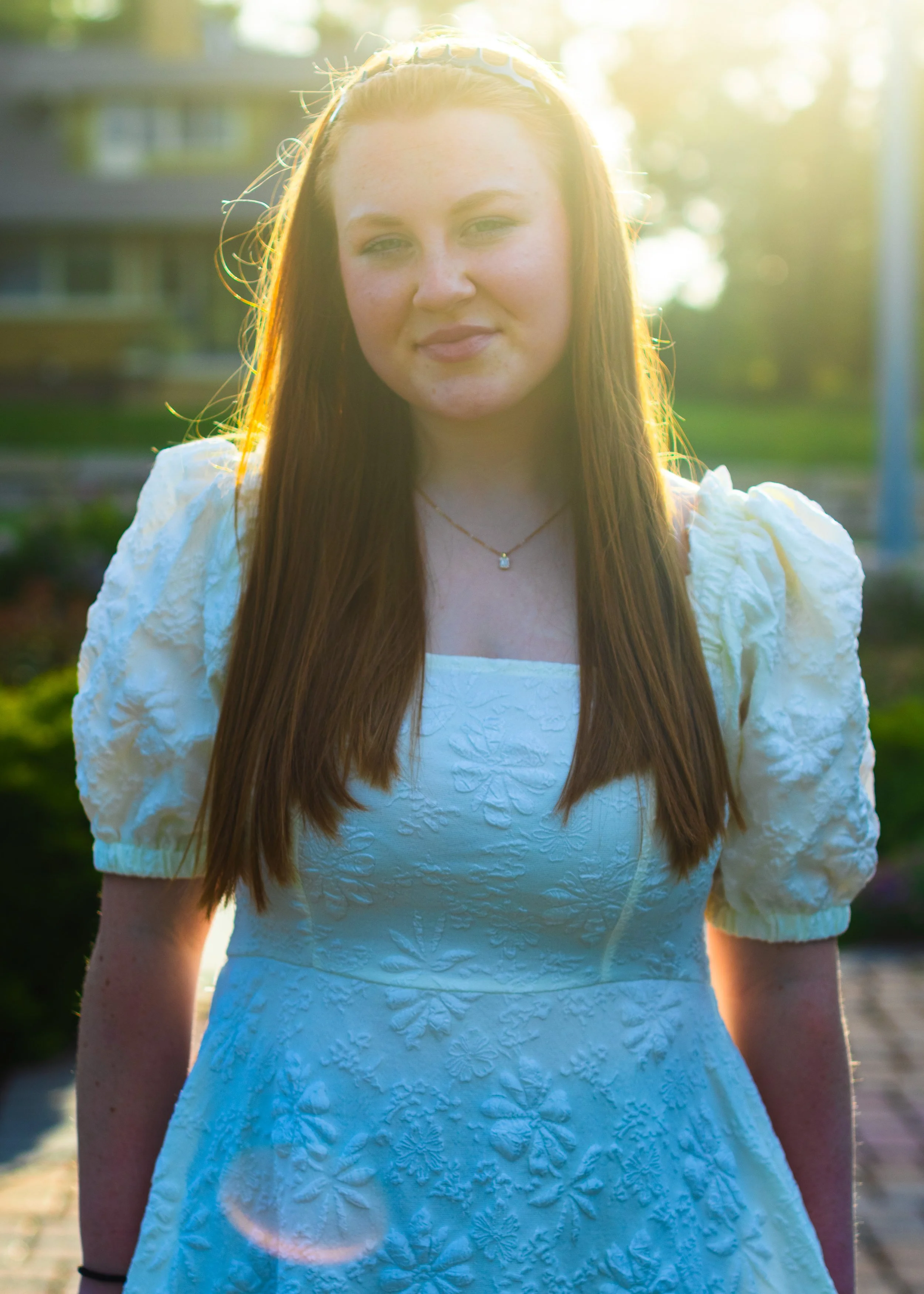 A young woman with long red hair wearing a white dress with puffed sleeves and a necklace, standing outdoors with sunlight and greenery in the background.