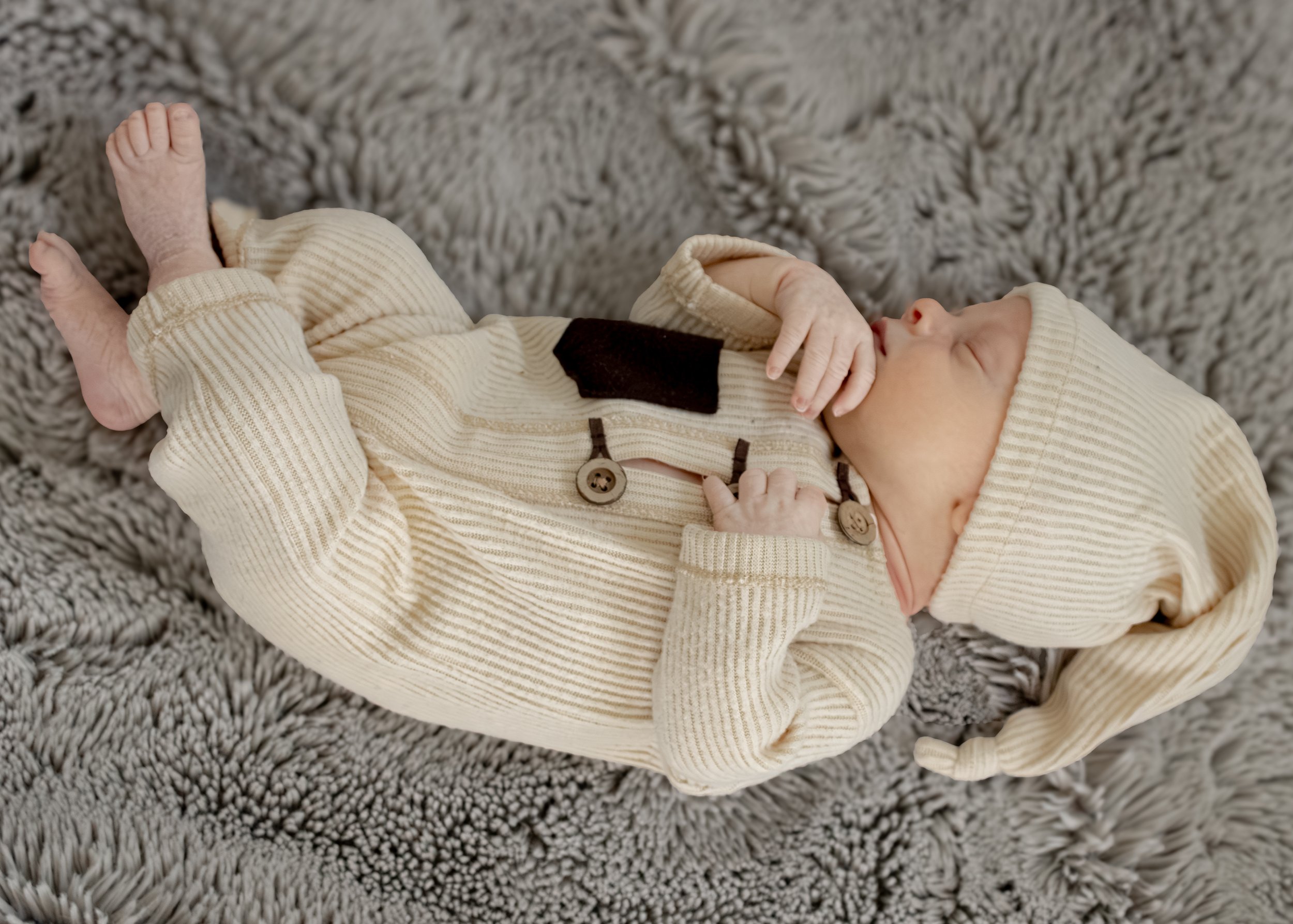 A newborn baby lying on a plush gray shaggy rug, wearing a cream-colored knitted sleeper and a matching hat.