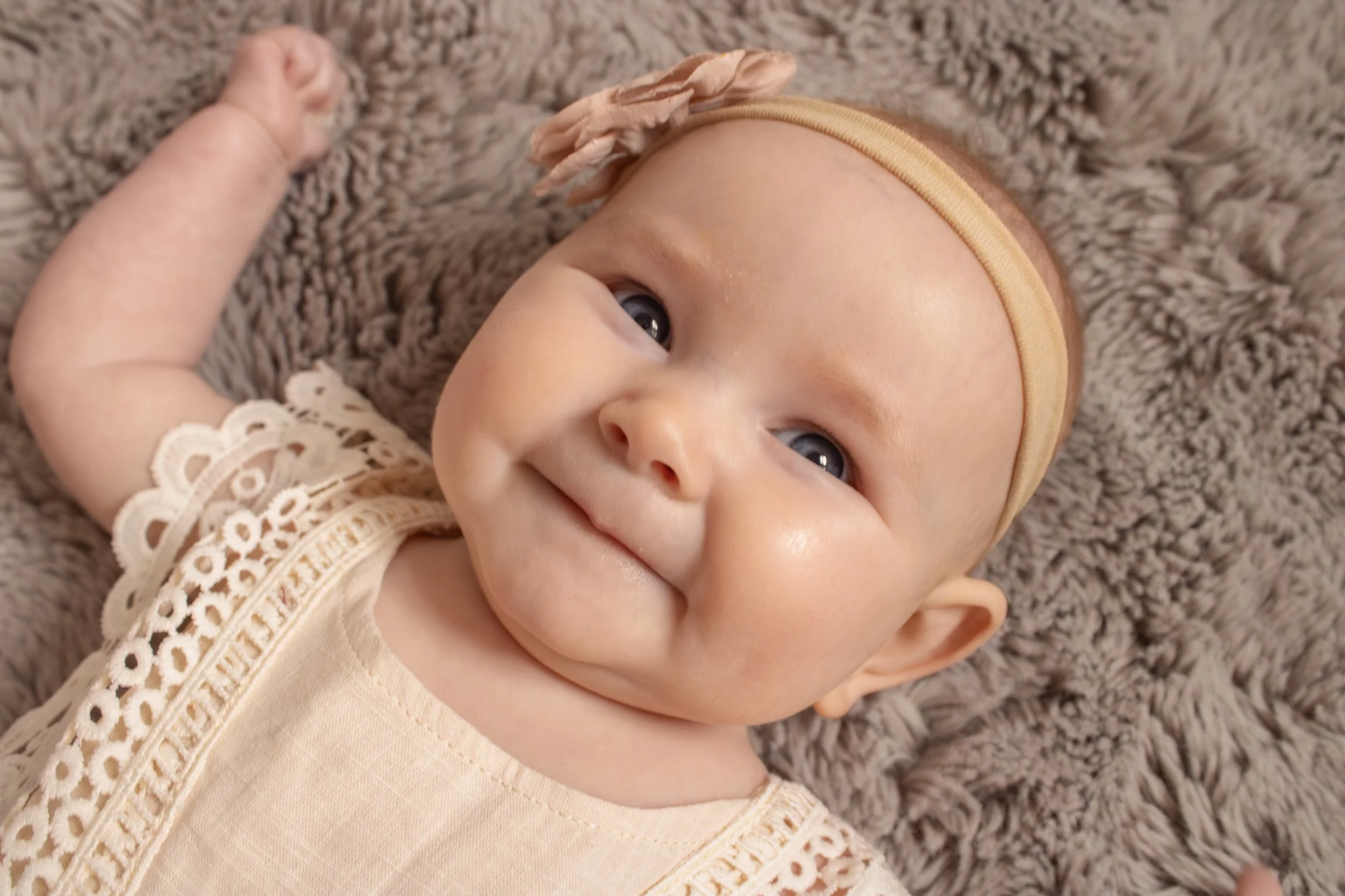 Close-up of a smiling baby lying on a soft, brown, furry blanket, wearing a beige dress with lace details and a beige headband with a flower decoration.