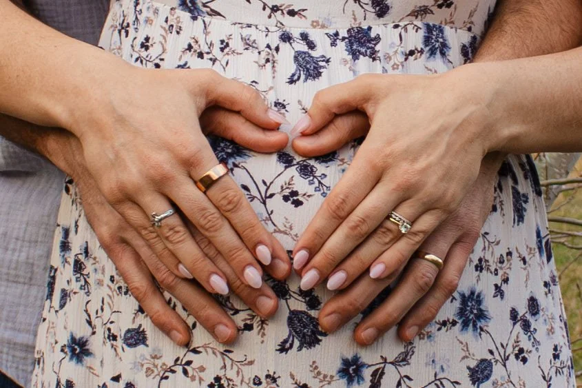 Multiple hands, including a pregnant woman's belly, forming a heart shape with their fingers, with wedding rings visible on some fingers.