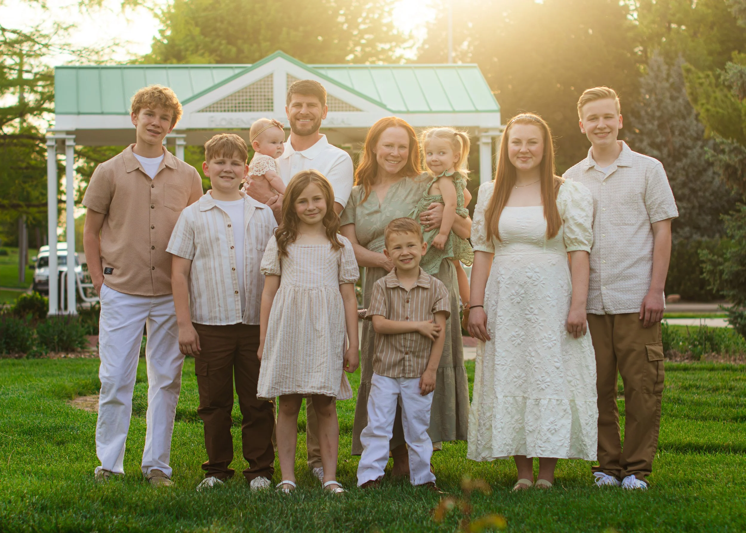 A multi-generational family standing together on a grassy area in front of a white gazebo with green roof, bathed in warm sunlight, smiling at the camera.