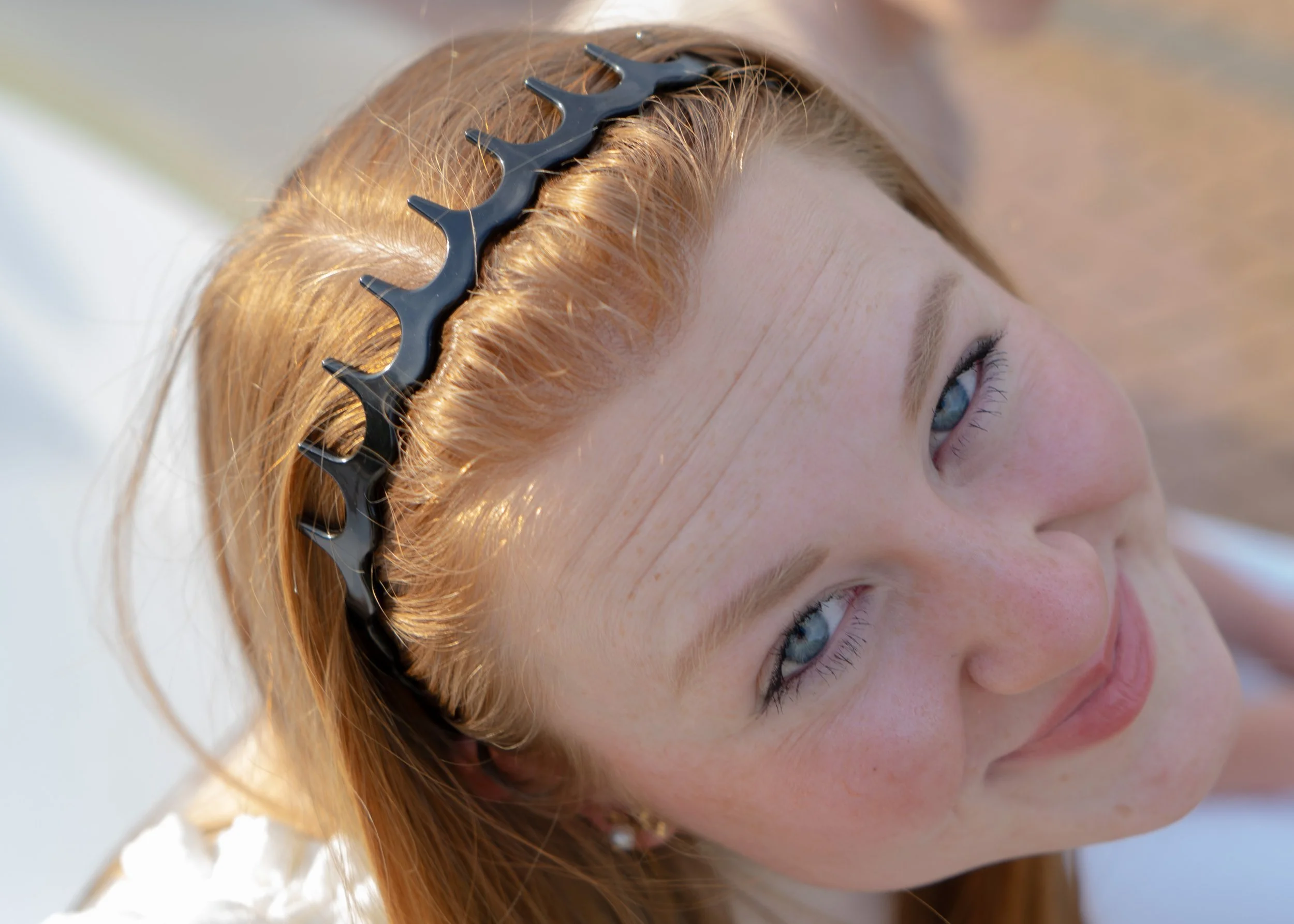 Close-up of a young woman with red hair, blue eyes, wearing a black hair clip, smiling, with a blurred background.