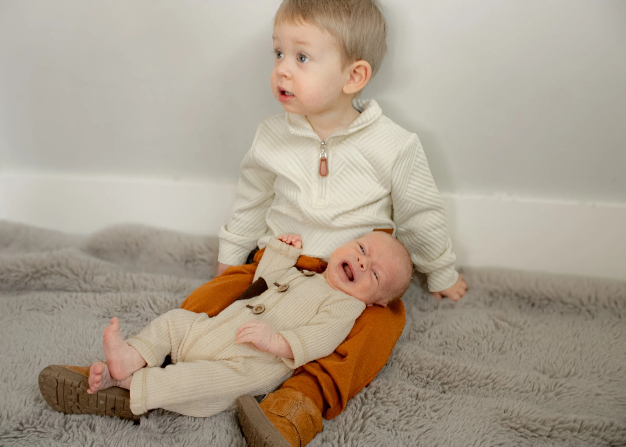 A young boy with light brown hair and a light-colored zip-up sweater sits on a gray shaggy rug, holding a newborn baby dressed in cream-colored clothing with brown accents. The boy looks to the side with his mouth slightly open, while the baby looks 