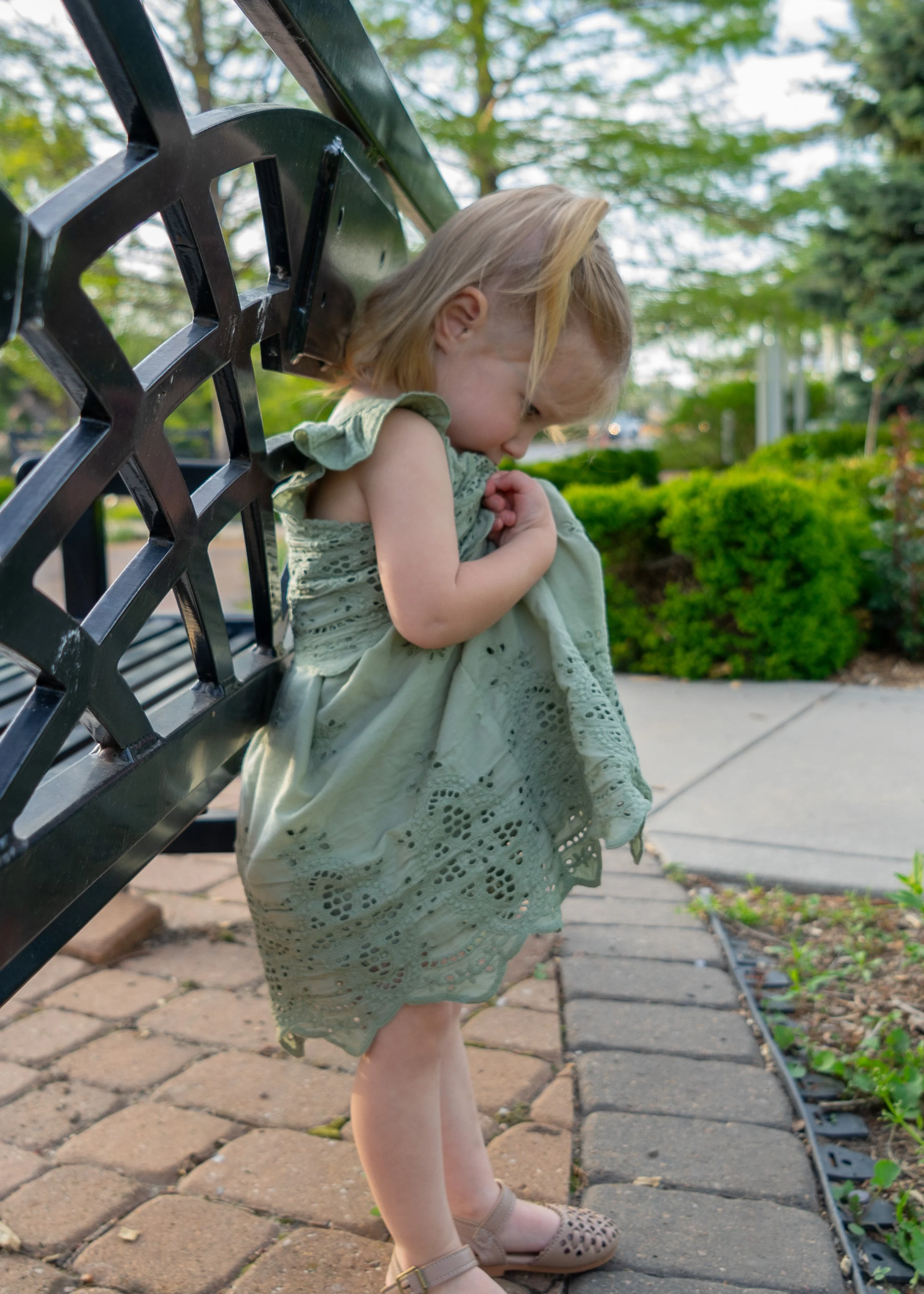 A young girl in a light green dress with eyelet details standing on a paved walkway beside a black metal park bench, holding her dress and looking down, with trees and bushes in the background.
