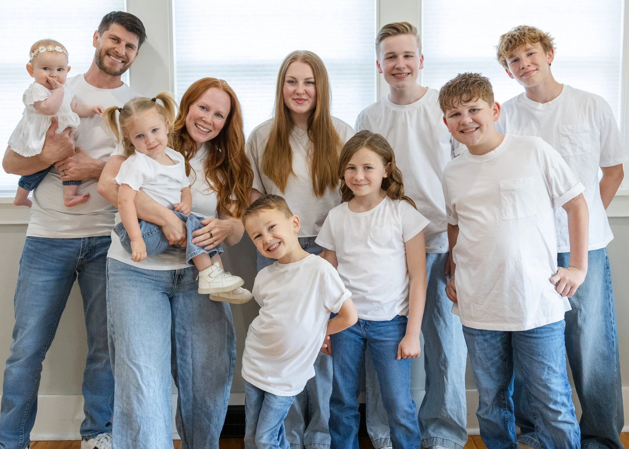 A diverse family group, including children and adults, standing together indoors in front of large windows wearing white shirts and blue jeans, smiling at the camera.