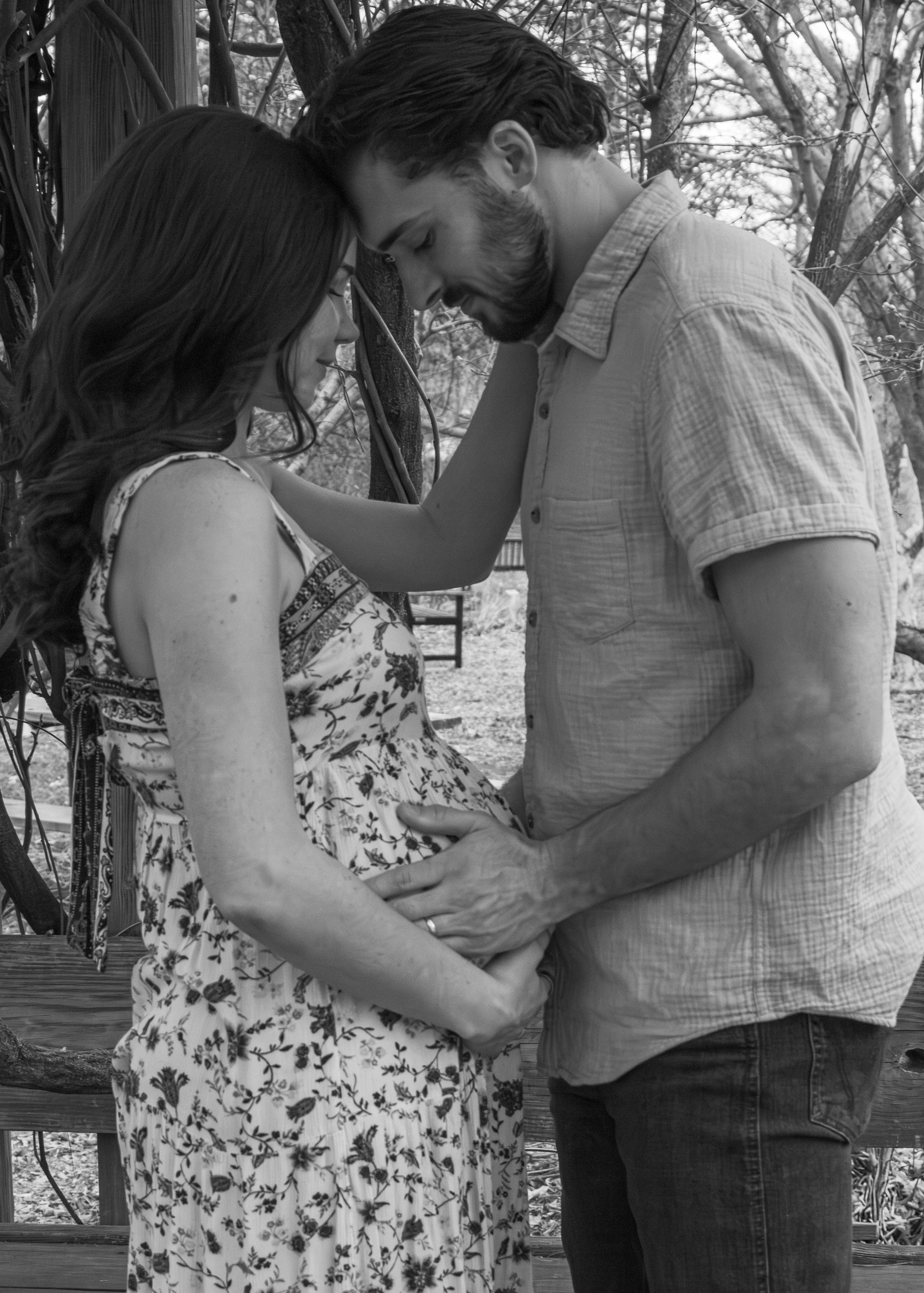 A couple stands close with foreheads touching, holding hands, in an outdoor setting surrounded by trees.