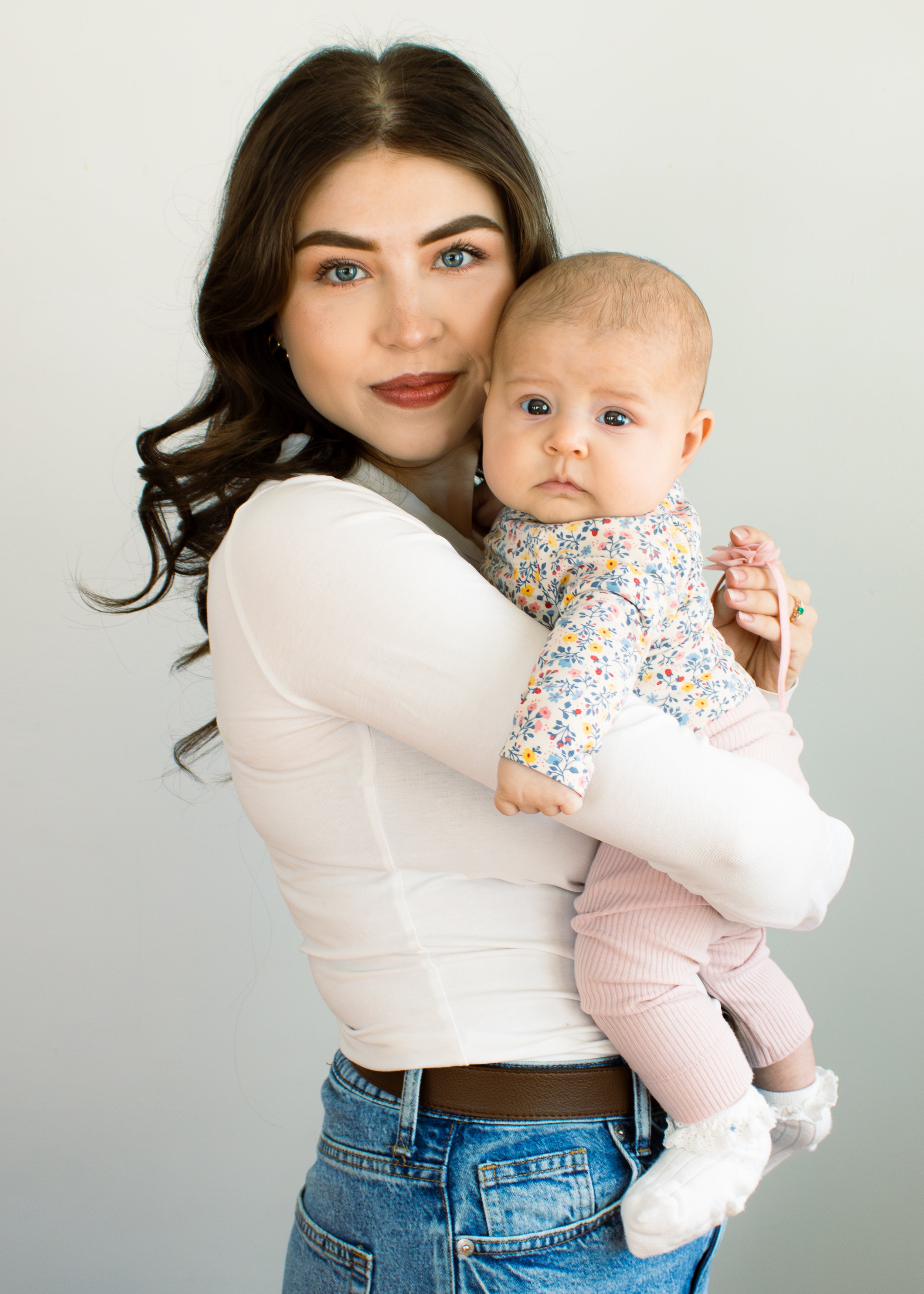 A woman with long wavy dark hair, blue eyes, and makeup, wearing a white shirt, holding a baby with light hair, bluish eyes, wearing a floral top and pink pants, against a plain light background.