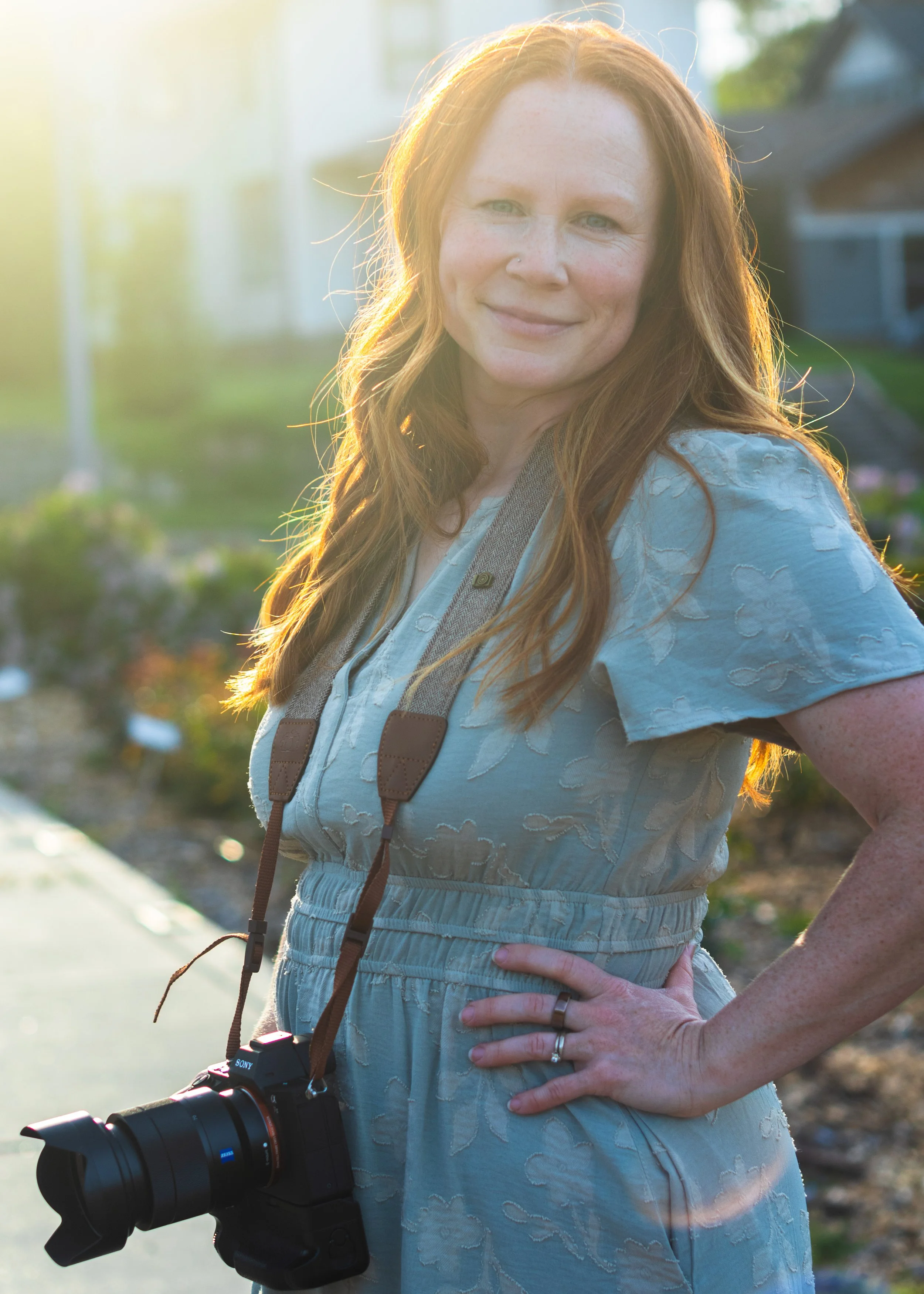 A woman with red hair and light skin, smiling outdoors during sunset, wearing a light blue dress with textured patterns and a camera hanging from her neck.