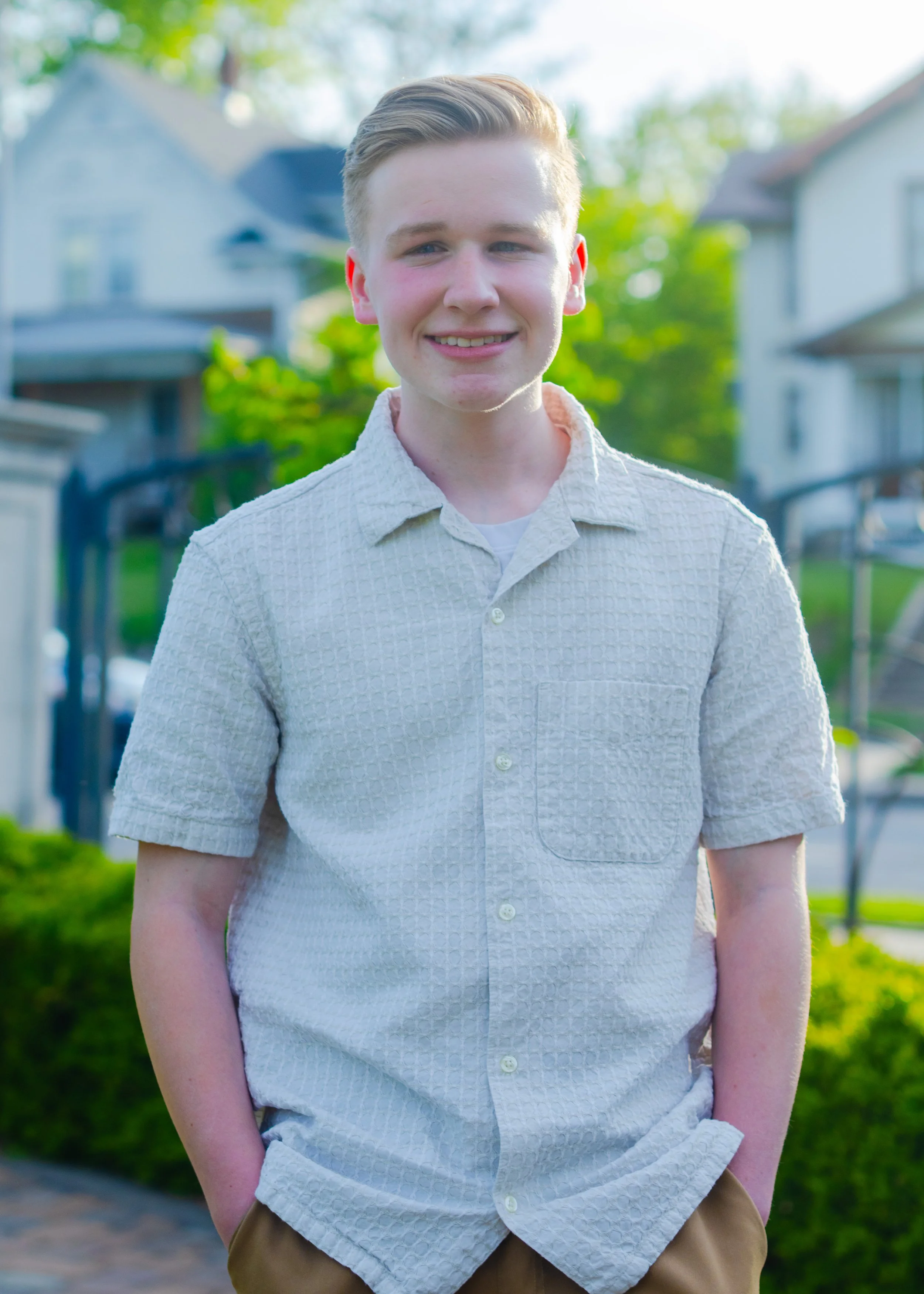 A young man with short, light brown hair standing outside in a residential neighborhood, smiling at the camera. He is wearing a light-colored, short-sleeved, button-up shirt and brown pants. 