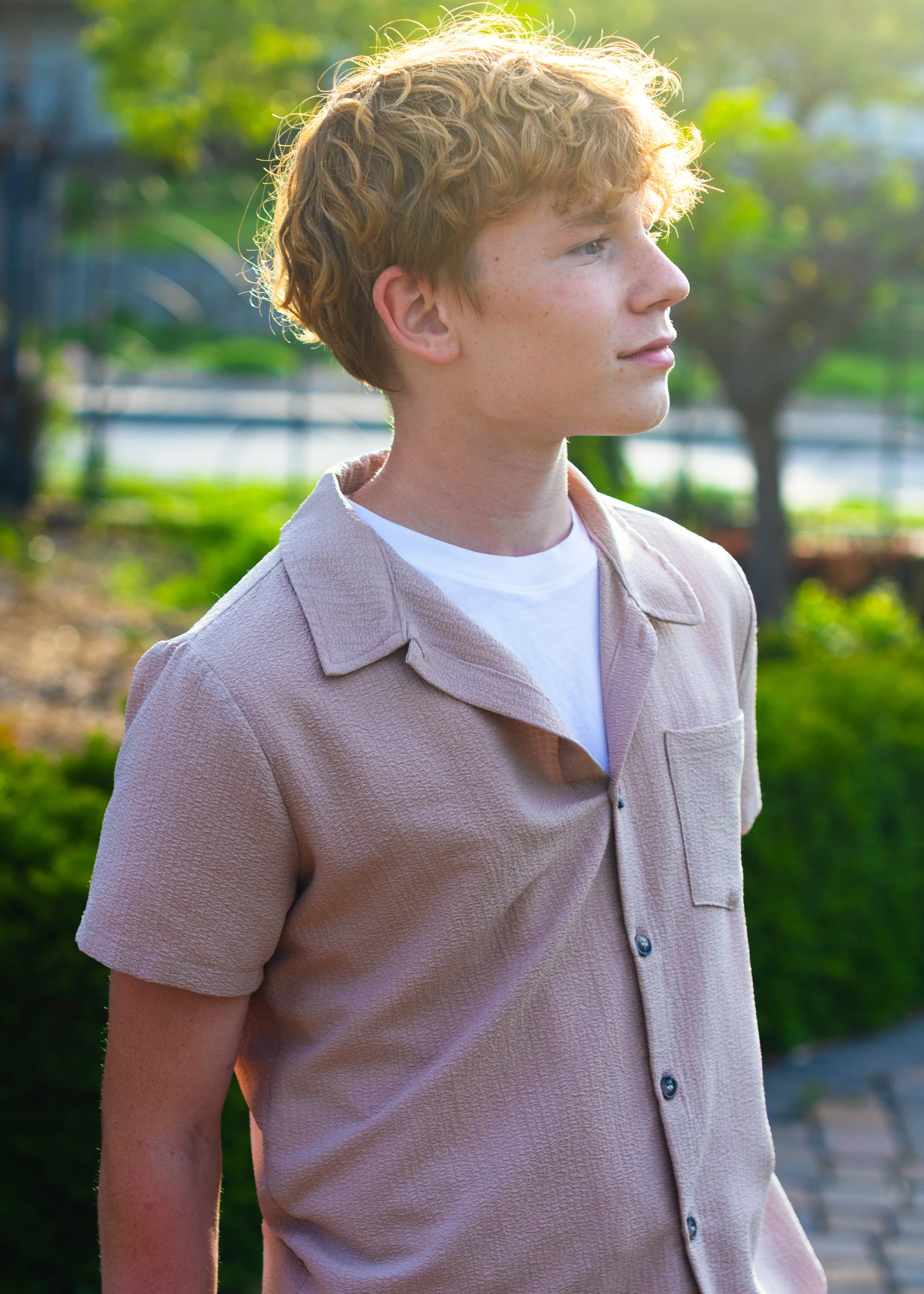 A young man with blond curly hair standing outdoors in a park on a sunny day, wearing a beige button-up shirt over a white t-shirt, looking to the right with a slight smile.