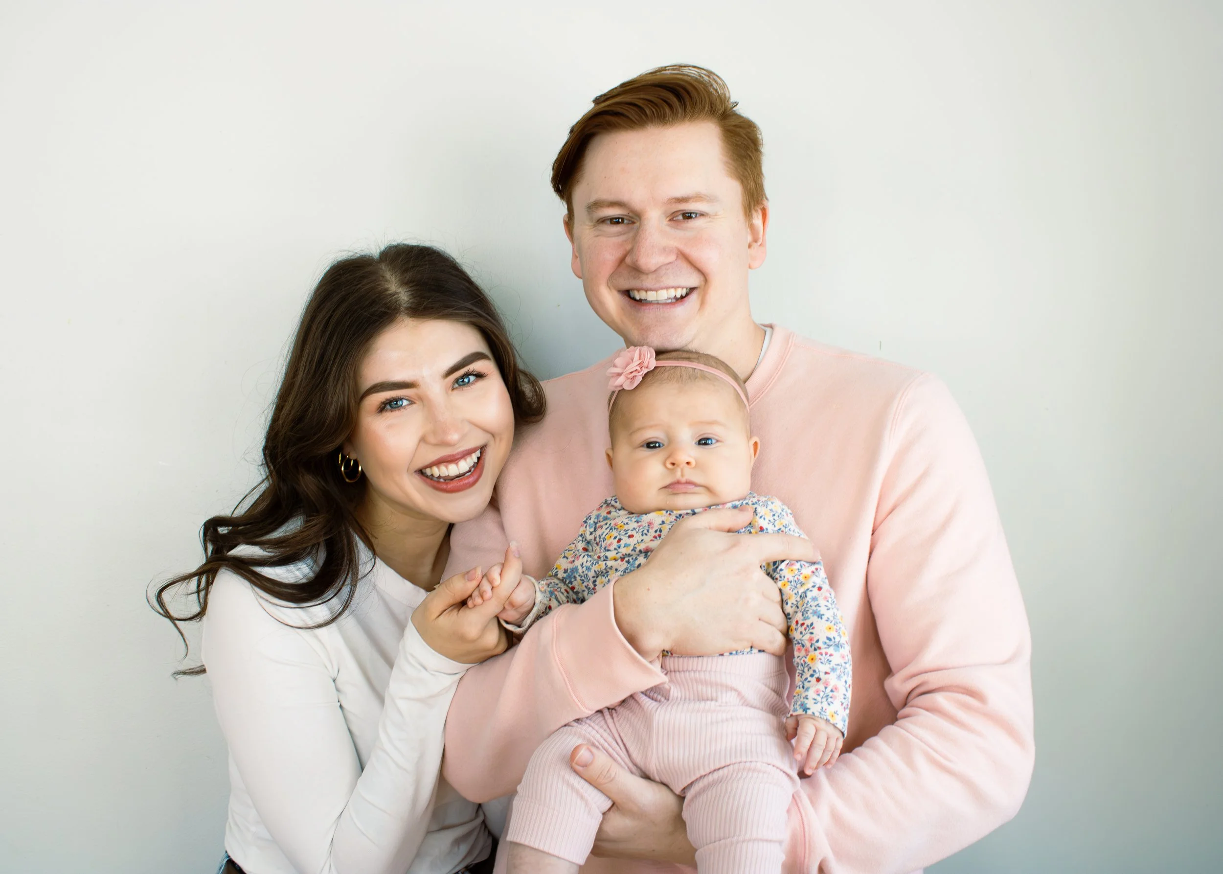 A happy family with a woman, a man, and a baby girl posing against a plain light-colored wall.