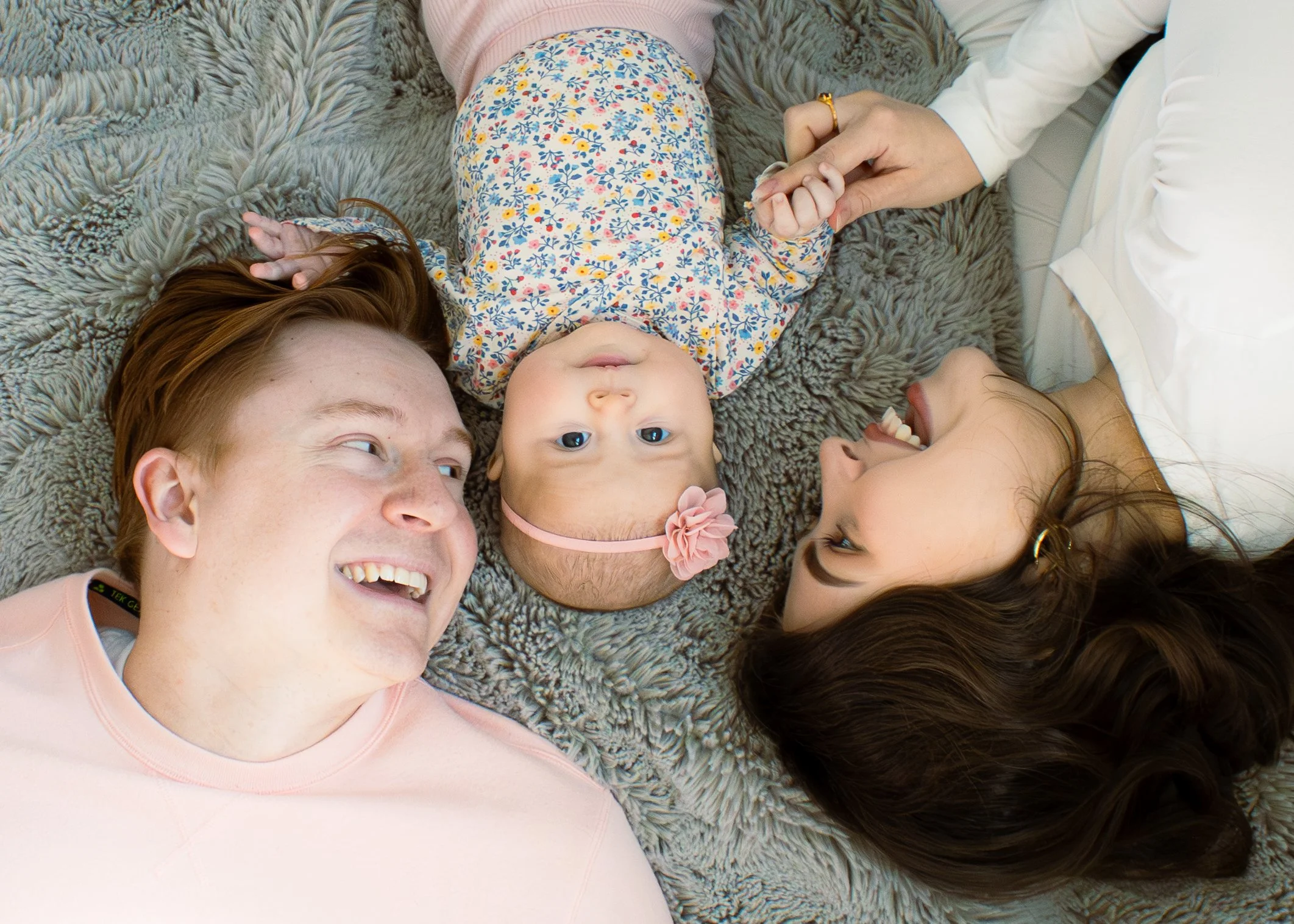 A family of four lying on a furry gray rug, holding hands, smiling, and looking at each other.