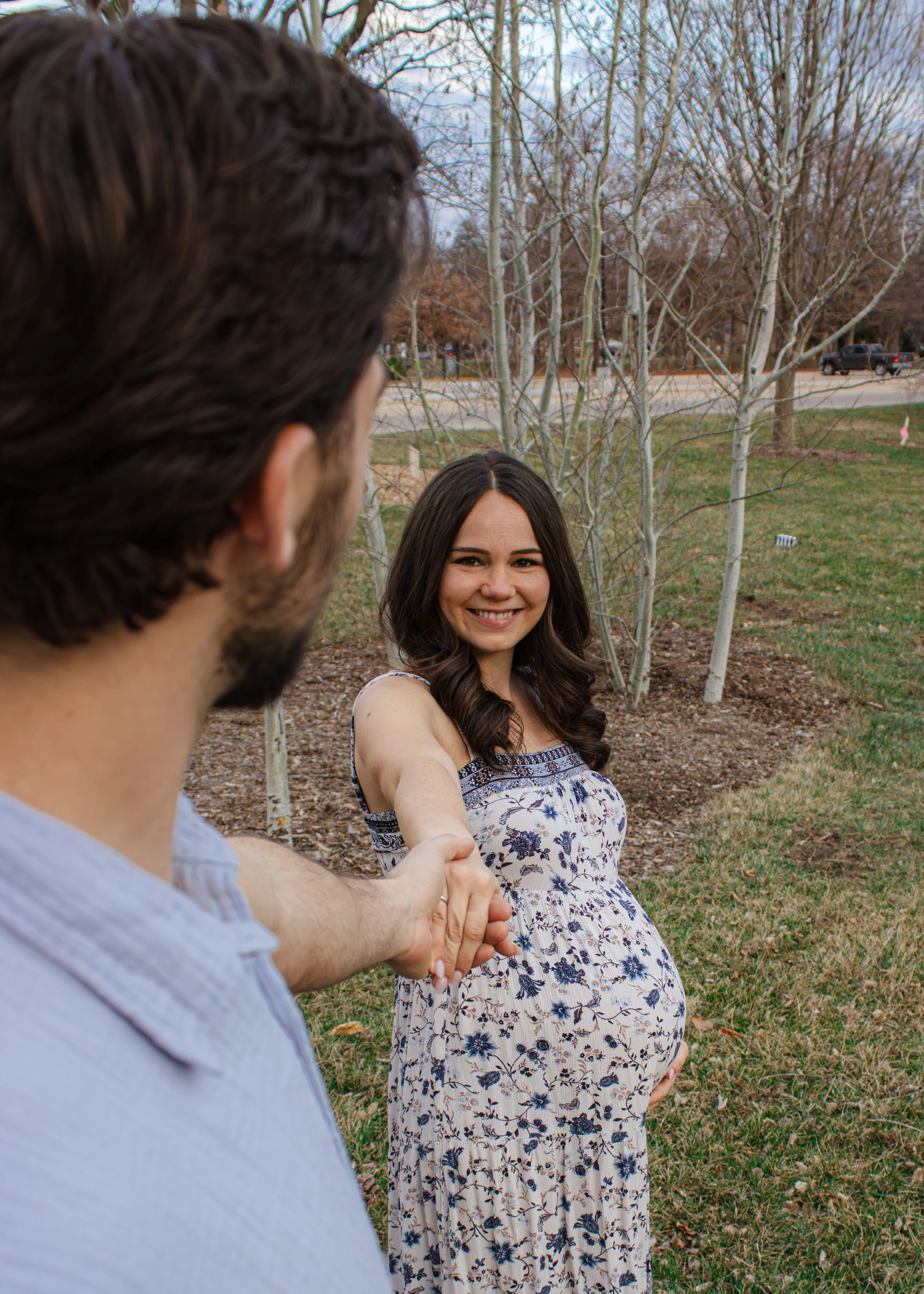 A woman in a floral dress smiling as she takes a selfie with a man whose face is out of focus, holding her hand, in a park with leafless trees and a grassy area.