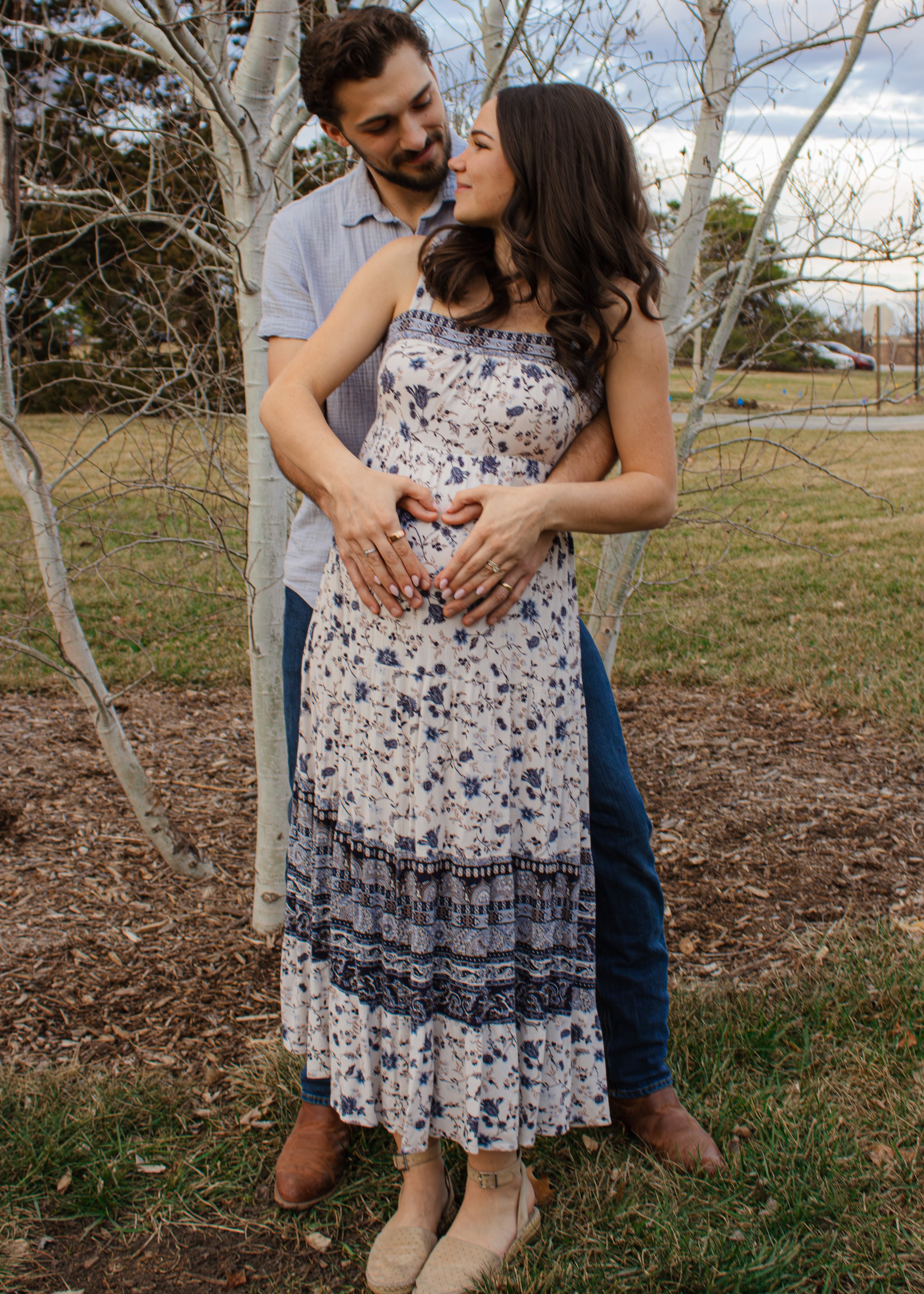 A couple standing outdoors under a leafless tree, with the man's arms around the woman's waist. The woman is pregnant, wearing a floral dress, and the man is dressed casually. They are looking at each other affectionately. The ground is covered with 