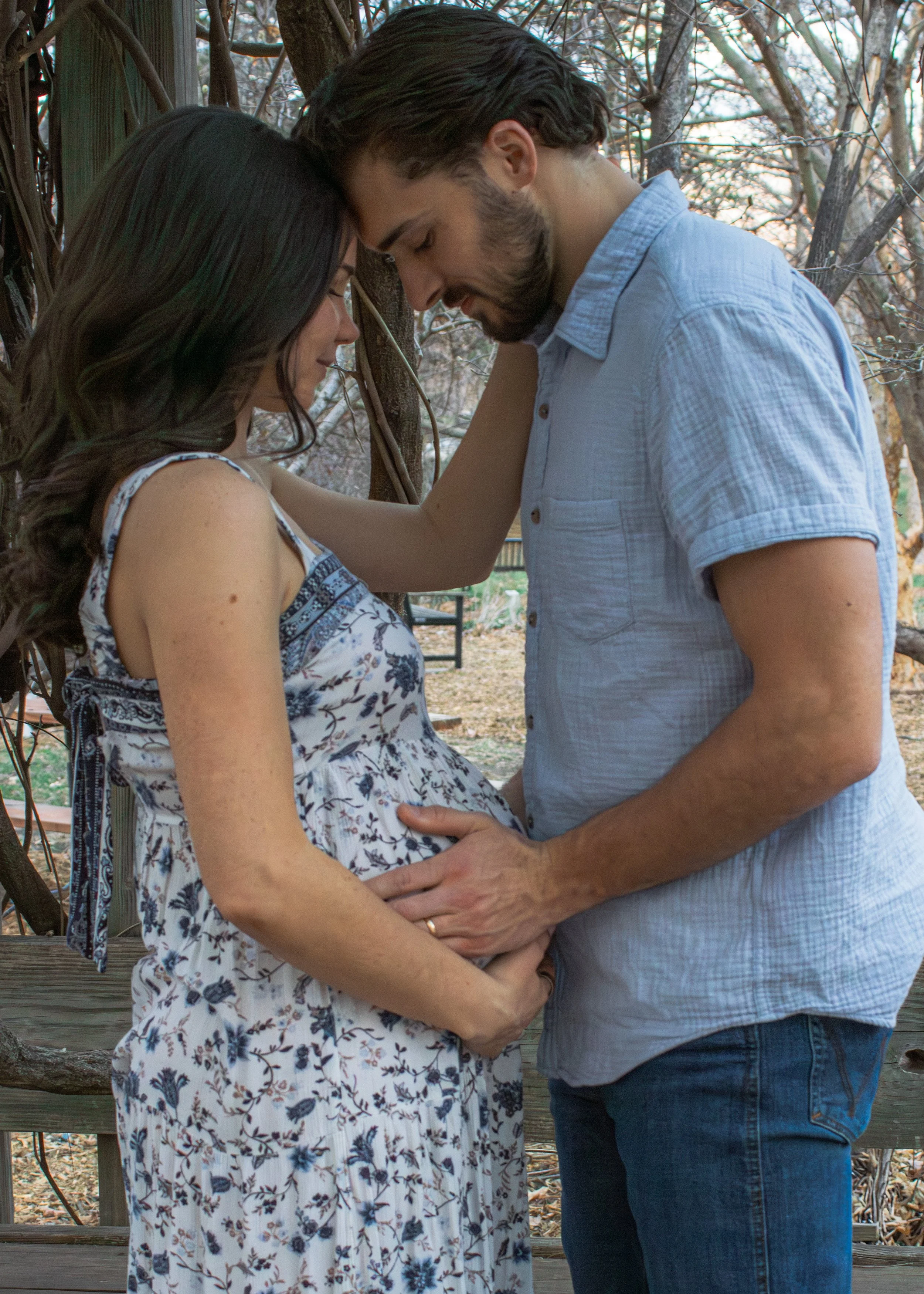A pregnant woman and a man standing close with their foreheads touching, holding her baby bump, outdoors near trees.
