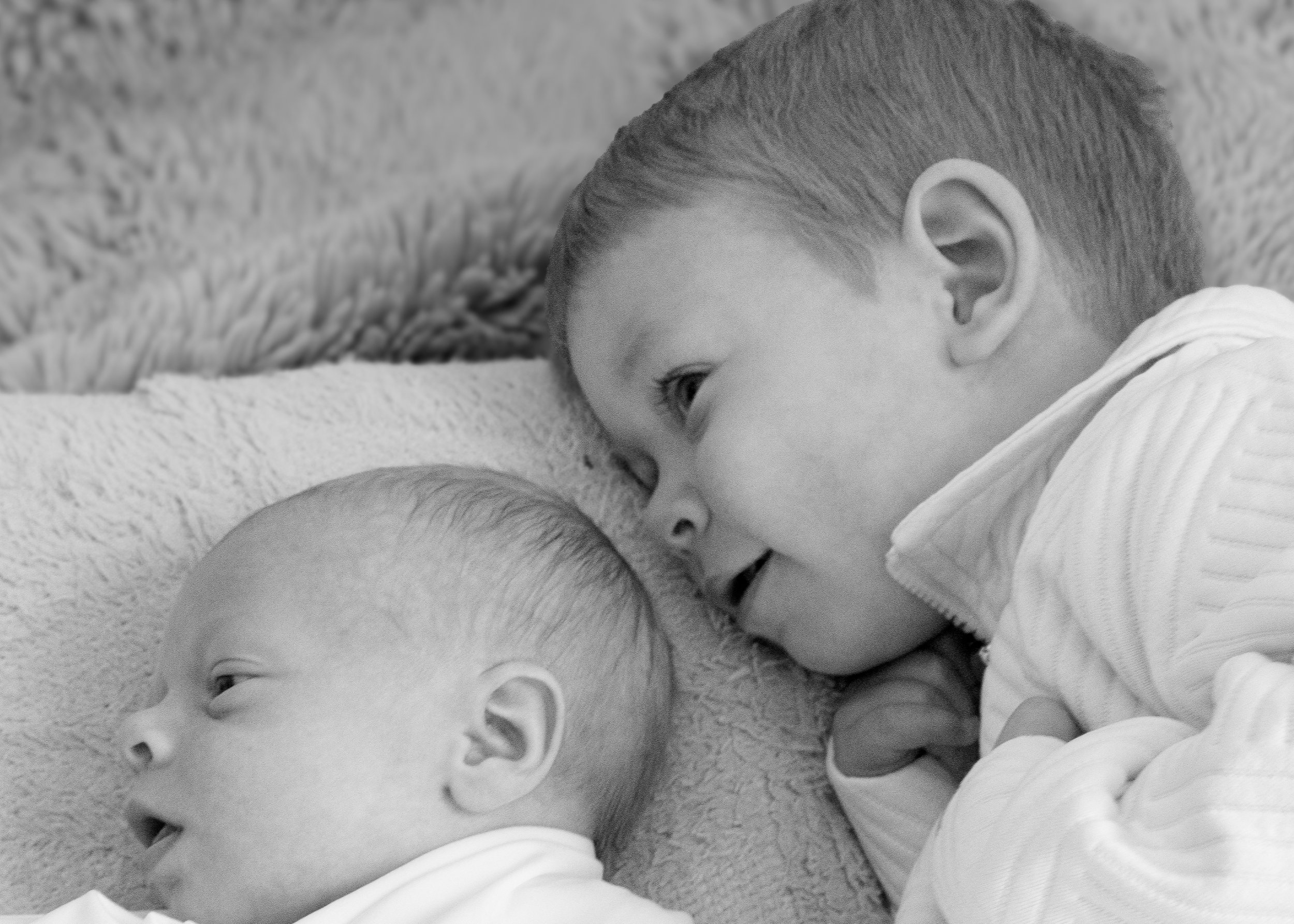 Close-up of two young children lying on the floor, facing each other, with neutral expressions, both wearing light-colored clothing.