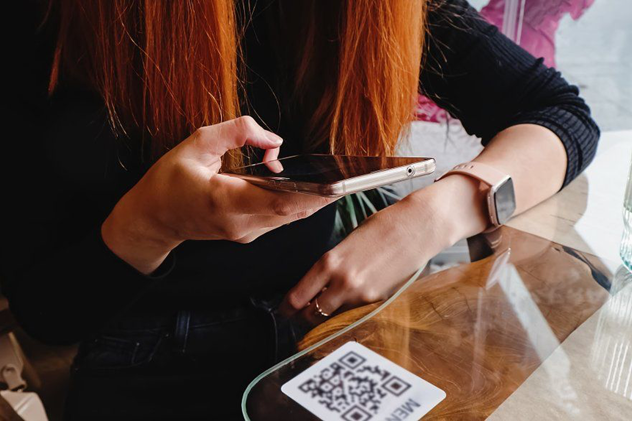 A woman with long red hair wearing a black long sleeve shirt and a smartwatch, sitting at a wooden table in a cafe, looking at her smartphone. There is a QR code sticker on the table and a water glass nearby.