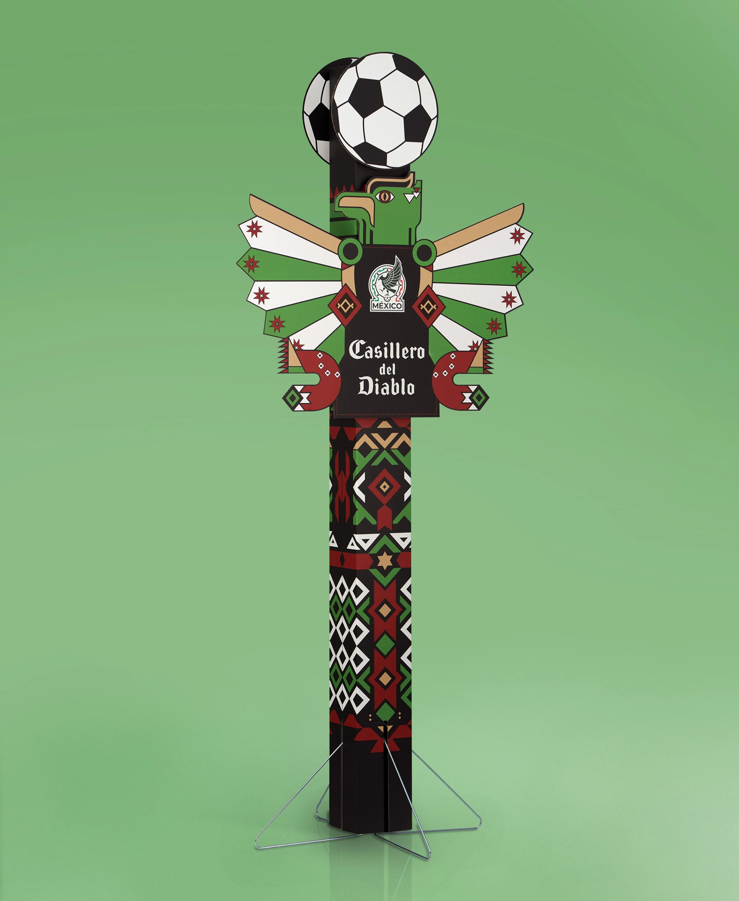 Decorative totem pole with a soccer ball on top, featuring vibrant geometric patterns, a crest with a wolf and the word 'México', and the phrase 'Casillero del Diablo' on the central part, standing on a plain green background.