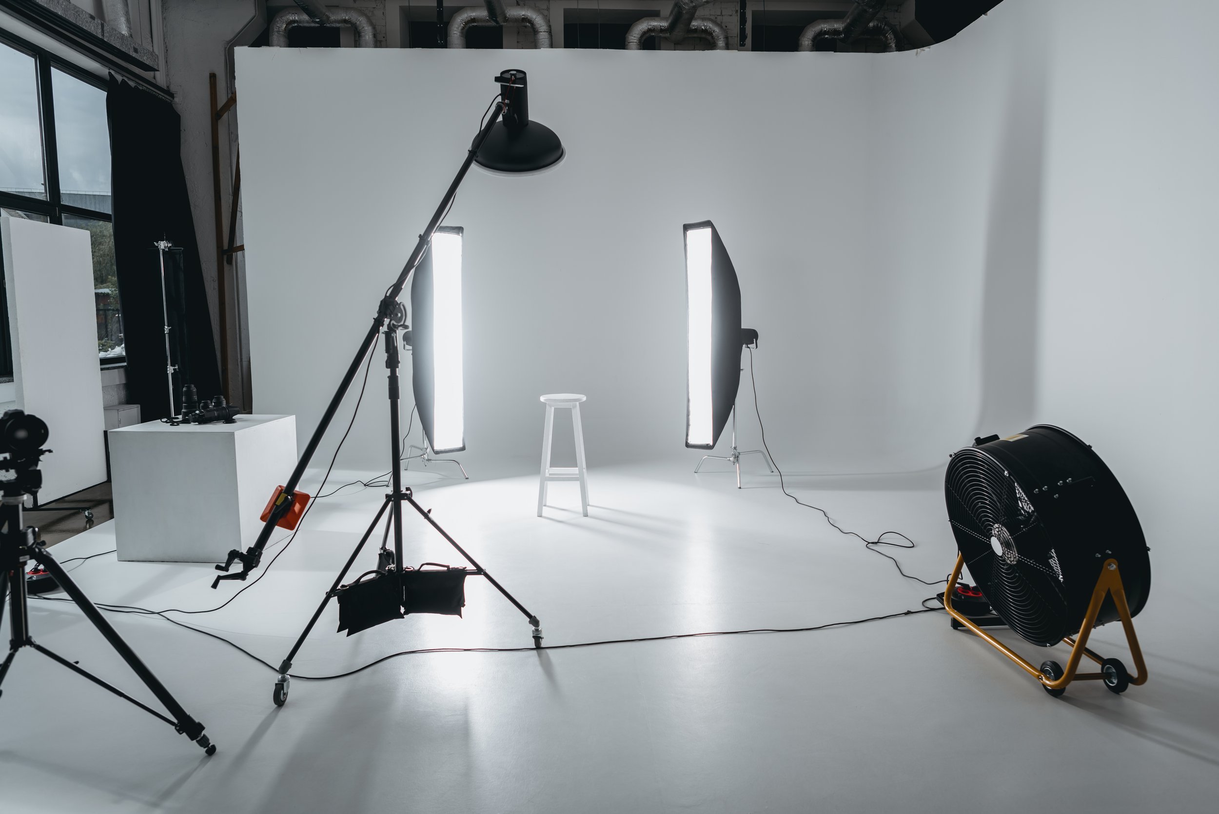 Photo of a photography studio with a white seamless backdrop, studio lights with softbox modifiers, a stool, multiple cameras and tripods, a fan, and other photography equipment.