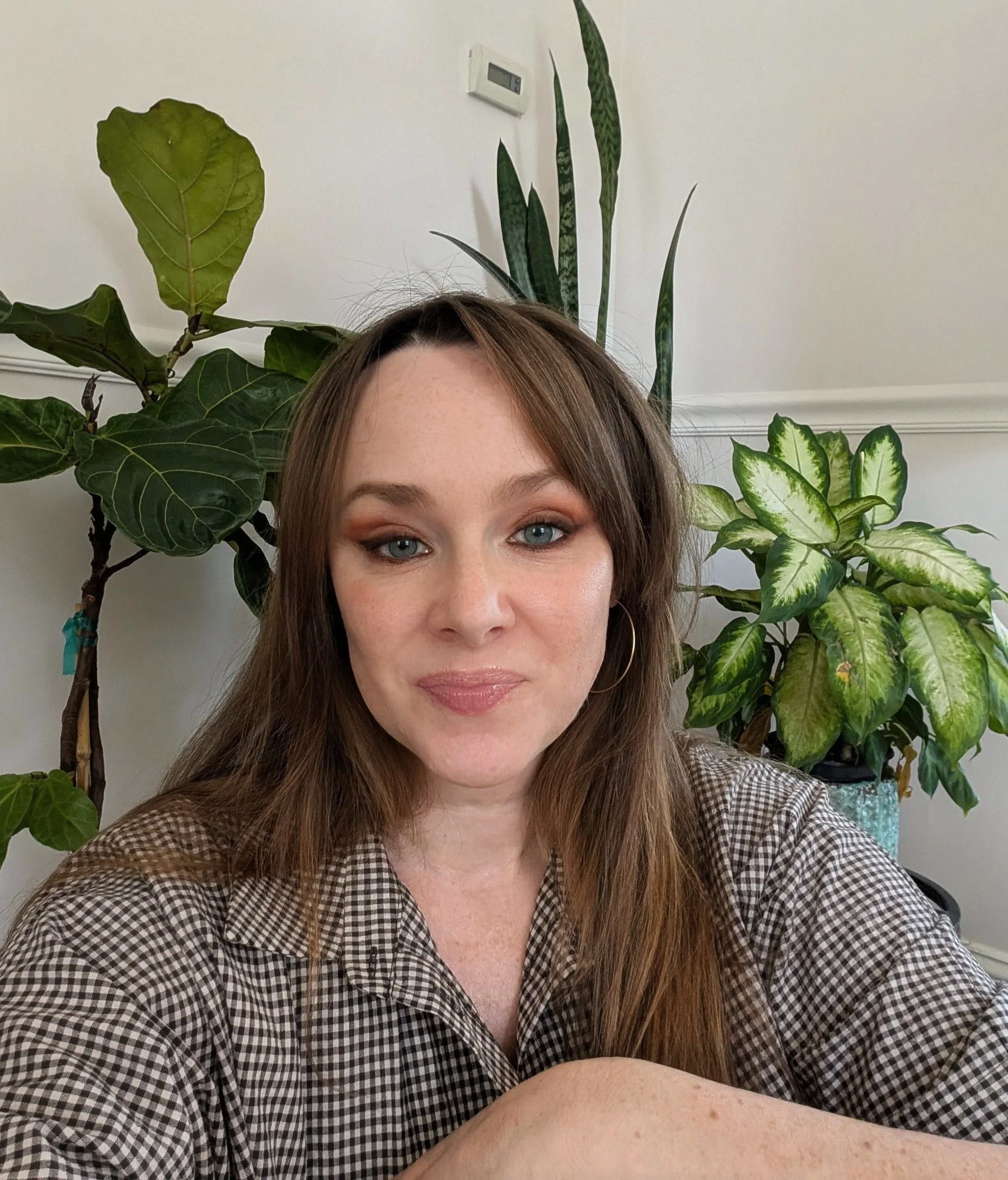 A woman with long brown hair and blue eyes sitting in front of large indoor green plants, wearing a checkered shirt and hoop earrings.