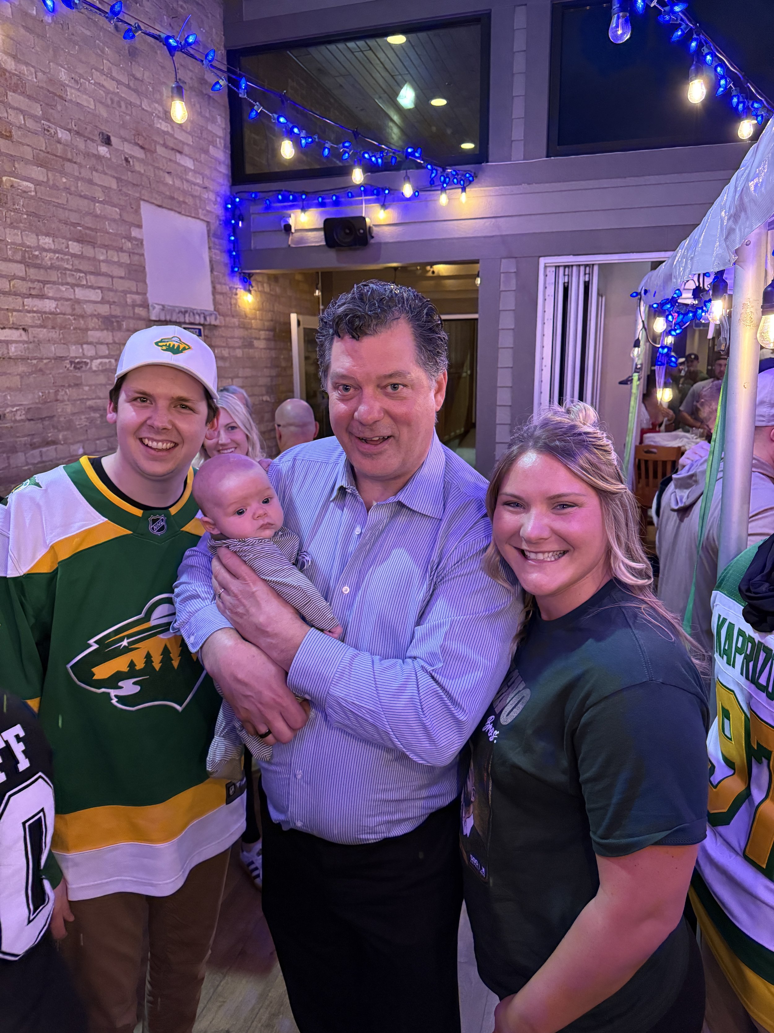 Group of people at a social gathering, including a man in a hockey jersey, a woman, a man holding a baby, and others, inside a venue decorated with colorful string lights.