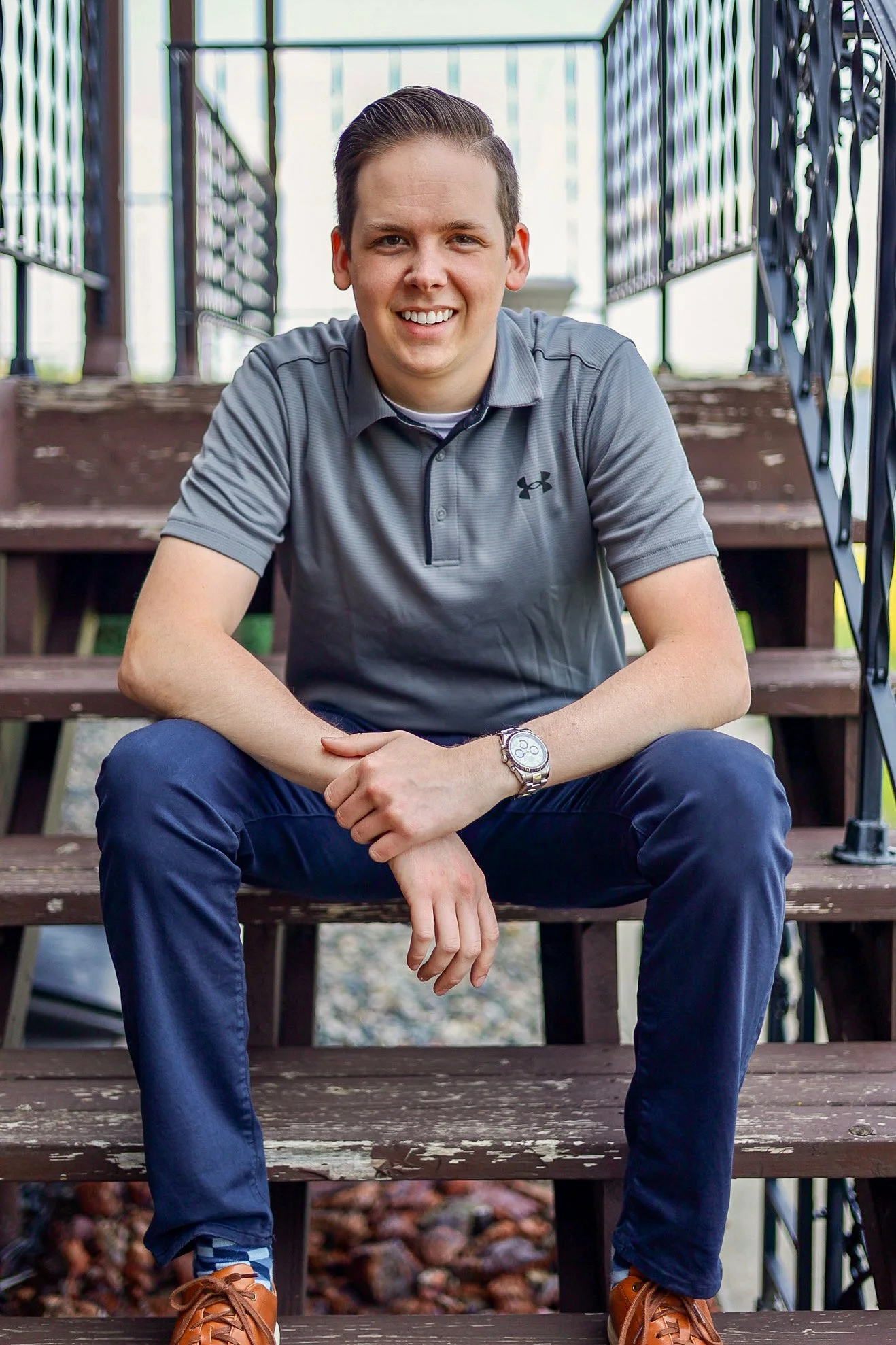 A young man with short brown hair, wearing a gray polo shirt and dark blue pants, is sitting on outdoor wooden stairs with black metal railings. He is smiling at the camera and has a watch on his left wrist. He is wearing brown shoes and colorful socks, and the background includes part of a brick pathway.