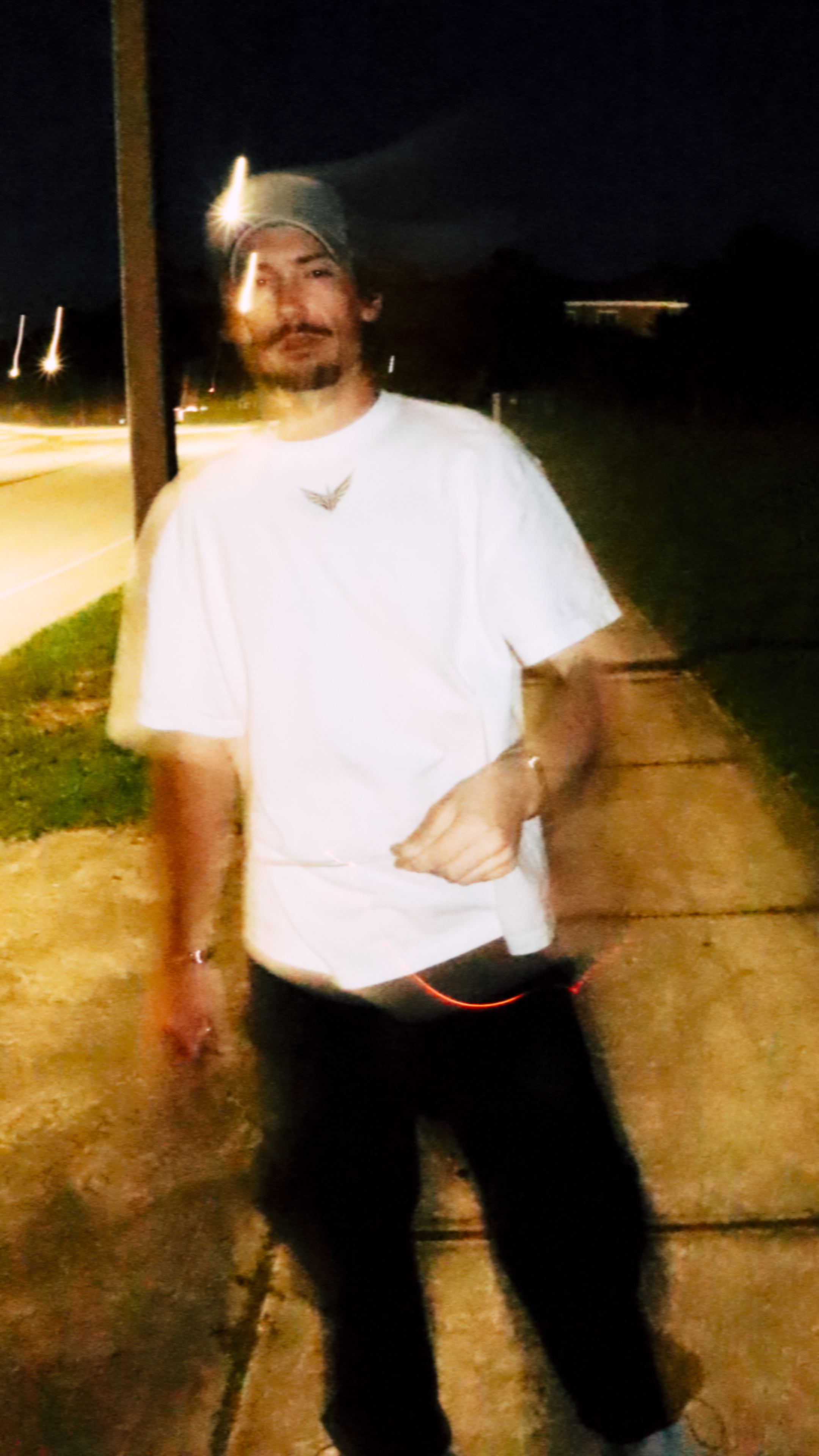 A man wearing a gray beanie, white t-shirt, and black pants standing outdoors at night on a sidewalk with streetlights in the background.