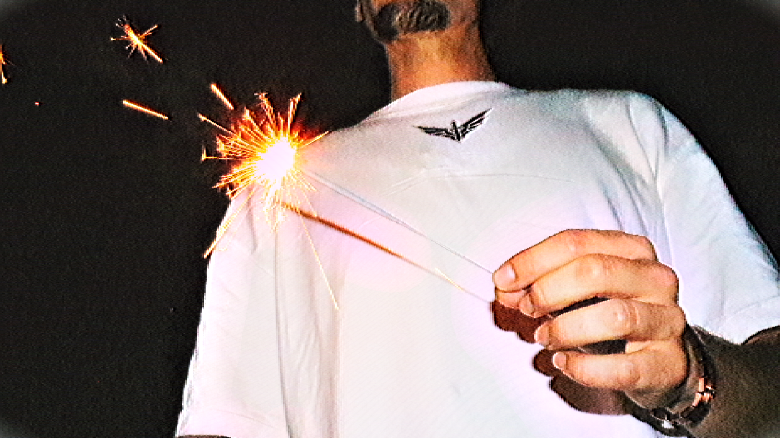 A person holding a lit sparkler in front of a black background, wearing a white shirt with a black emblem on the chest.