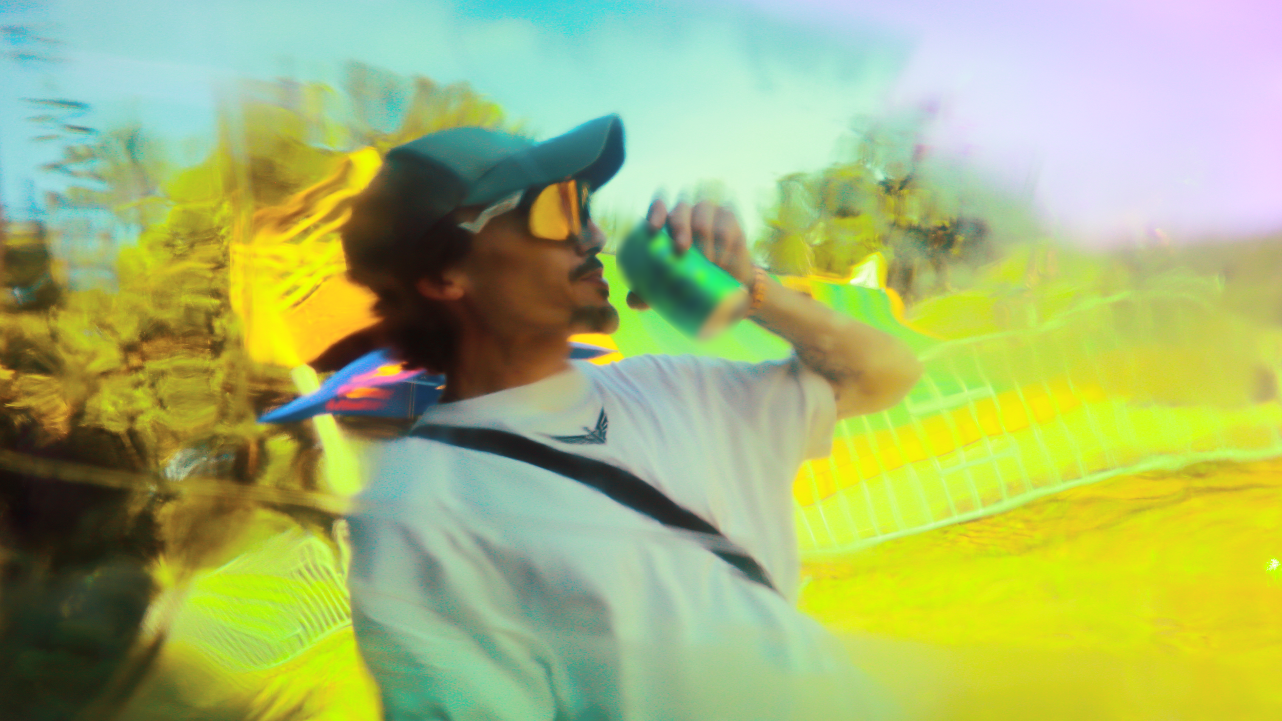 Person wearing sunglasses and a cap, holding a green bottle, with a blurred colorful background.