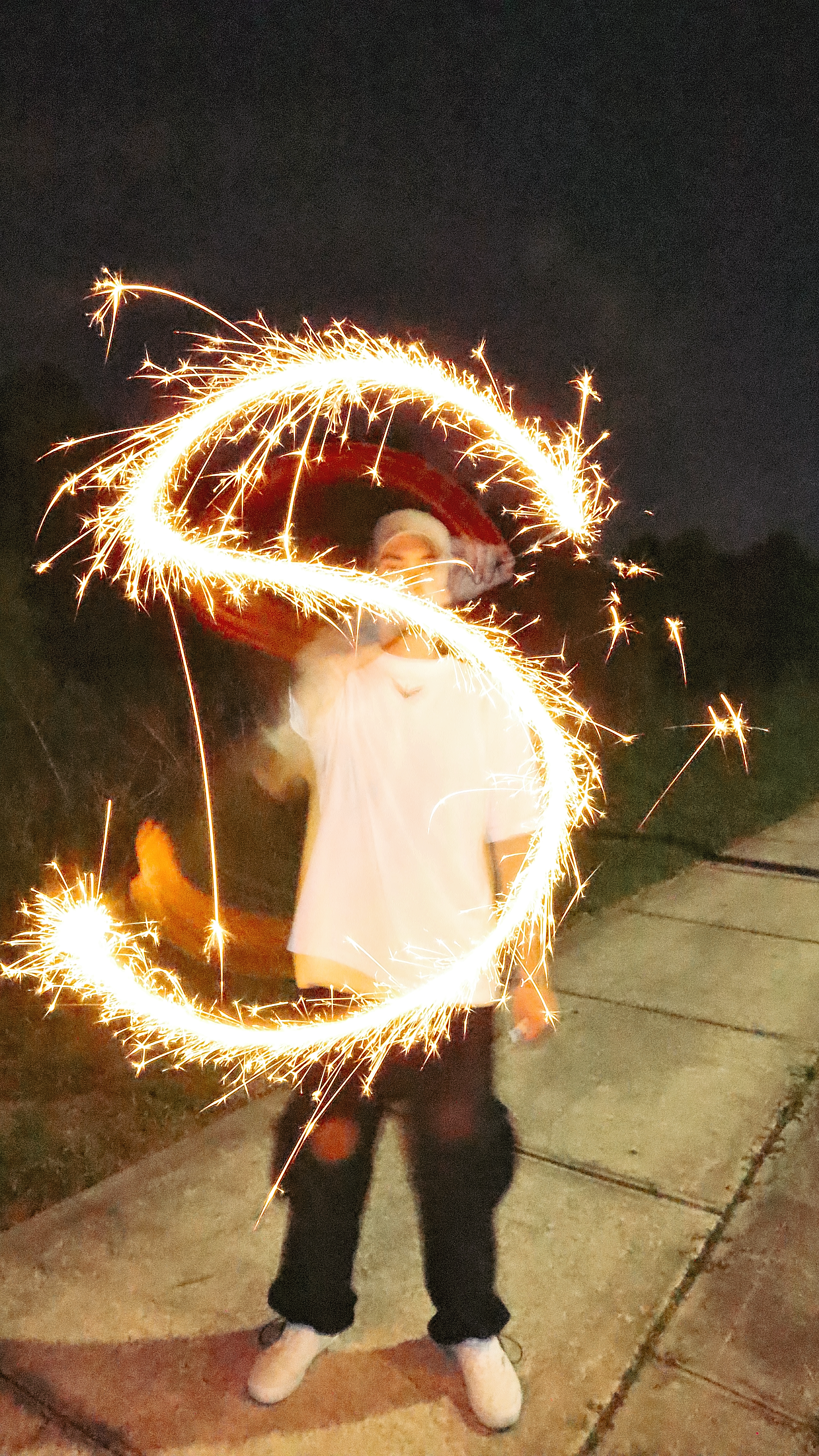 A person wearing a Santa hat and white shirt is playing with sparklers, creating a circular spark pattern at night outdoors.