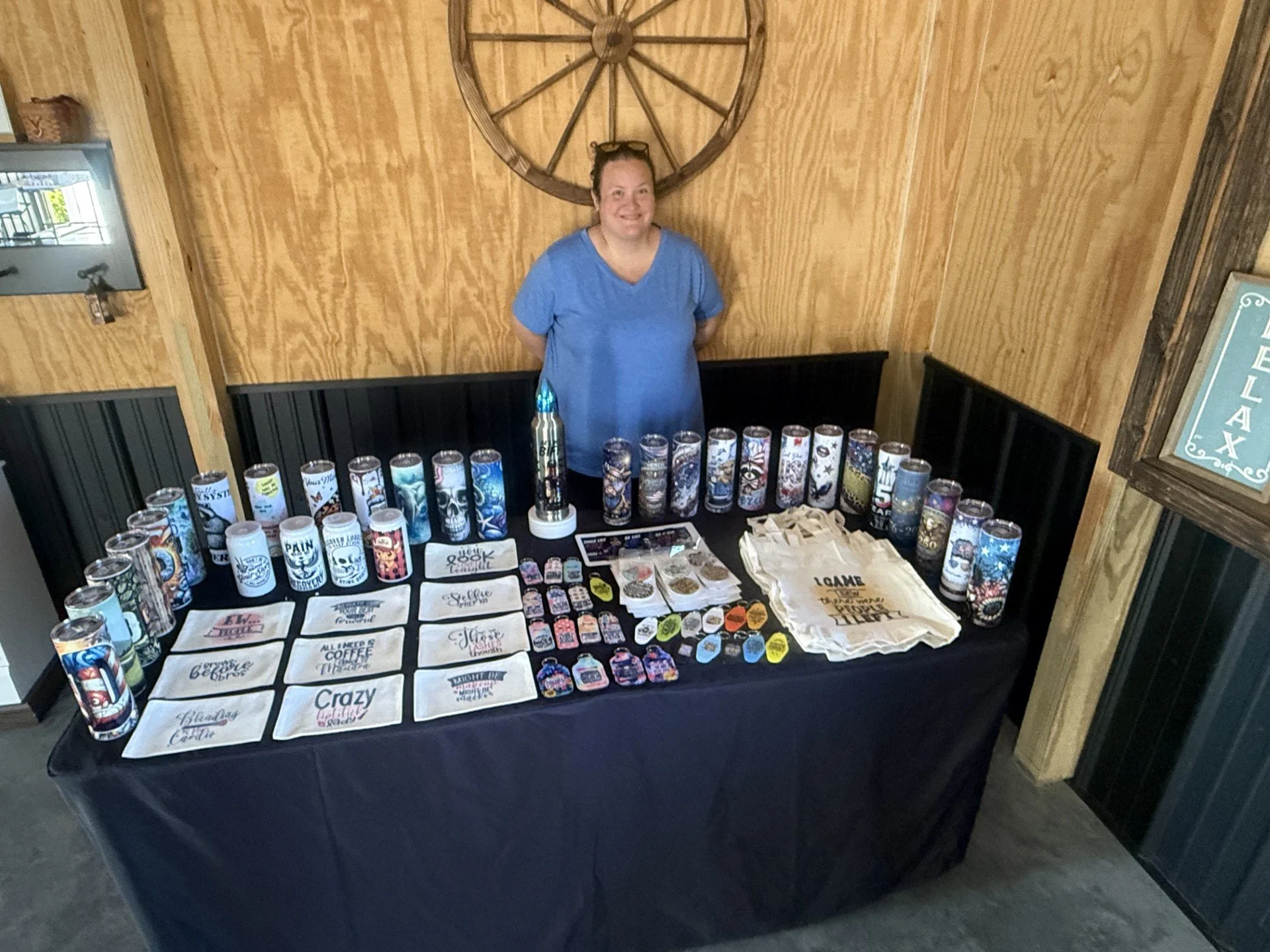 A woman standing behind a display table with various cups, small items, and tote bags with text and images, in a rustic wood-paneled room.