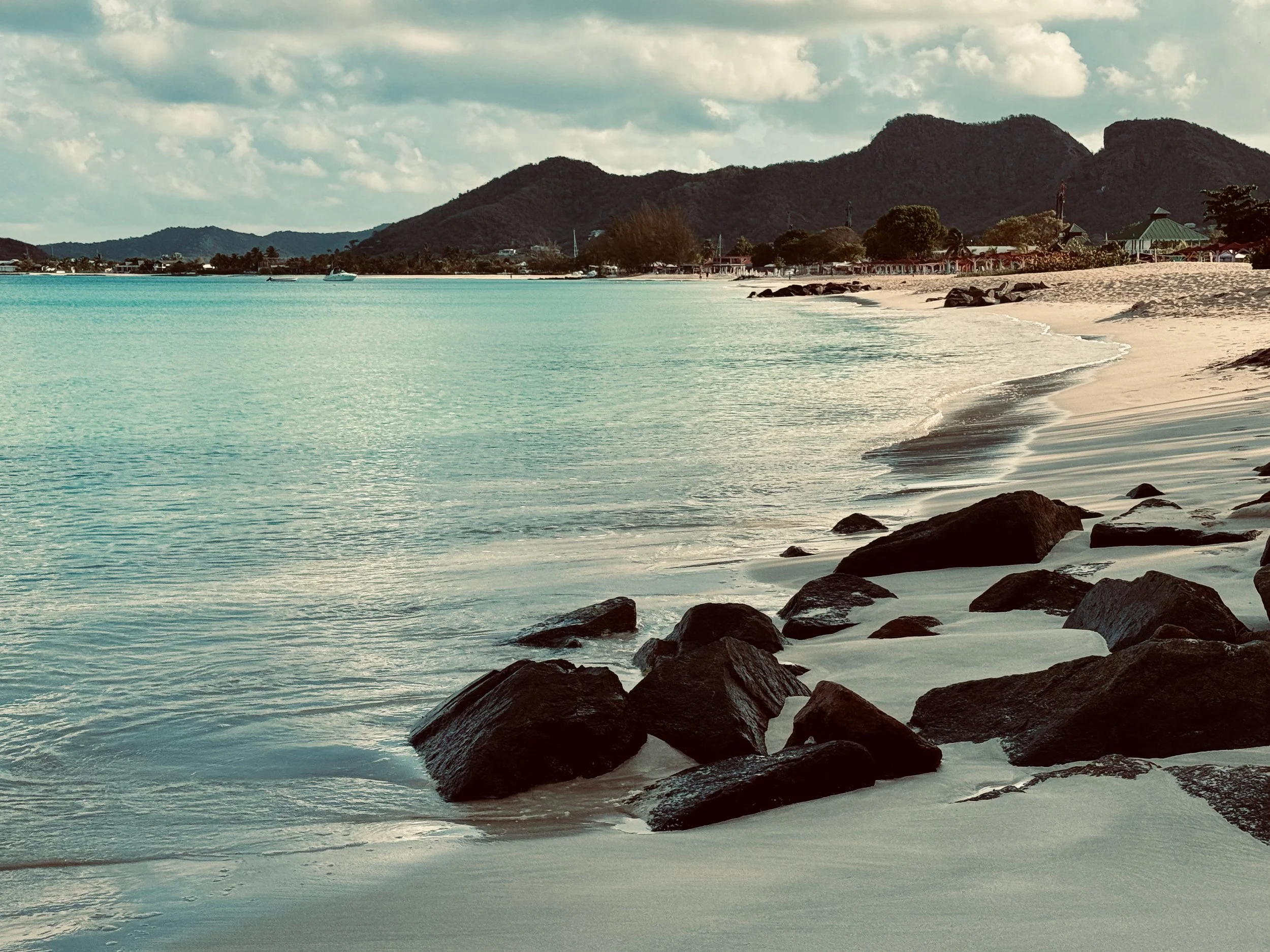 A scenic beach with calm turquoise water, large rocks along the shoreline, and a sandy beach area with buildings and trees. Mountain ranges are visible in the background under a partly cloudy sky.