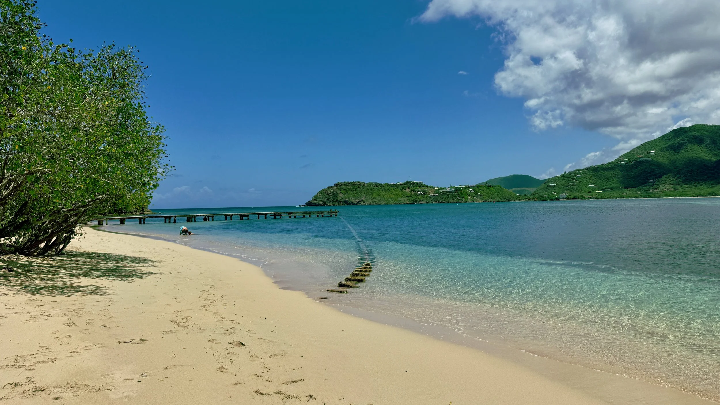 A beach with white sand, clear blue water, green hills in the background, and a partly cloudy sky.