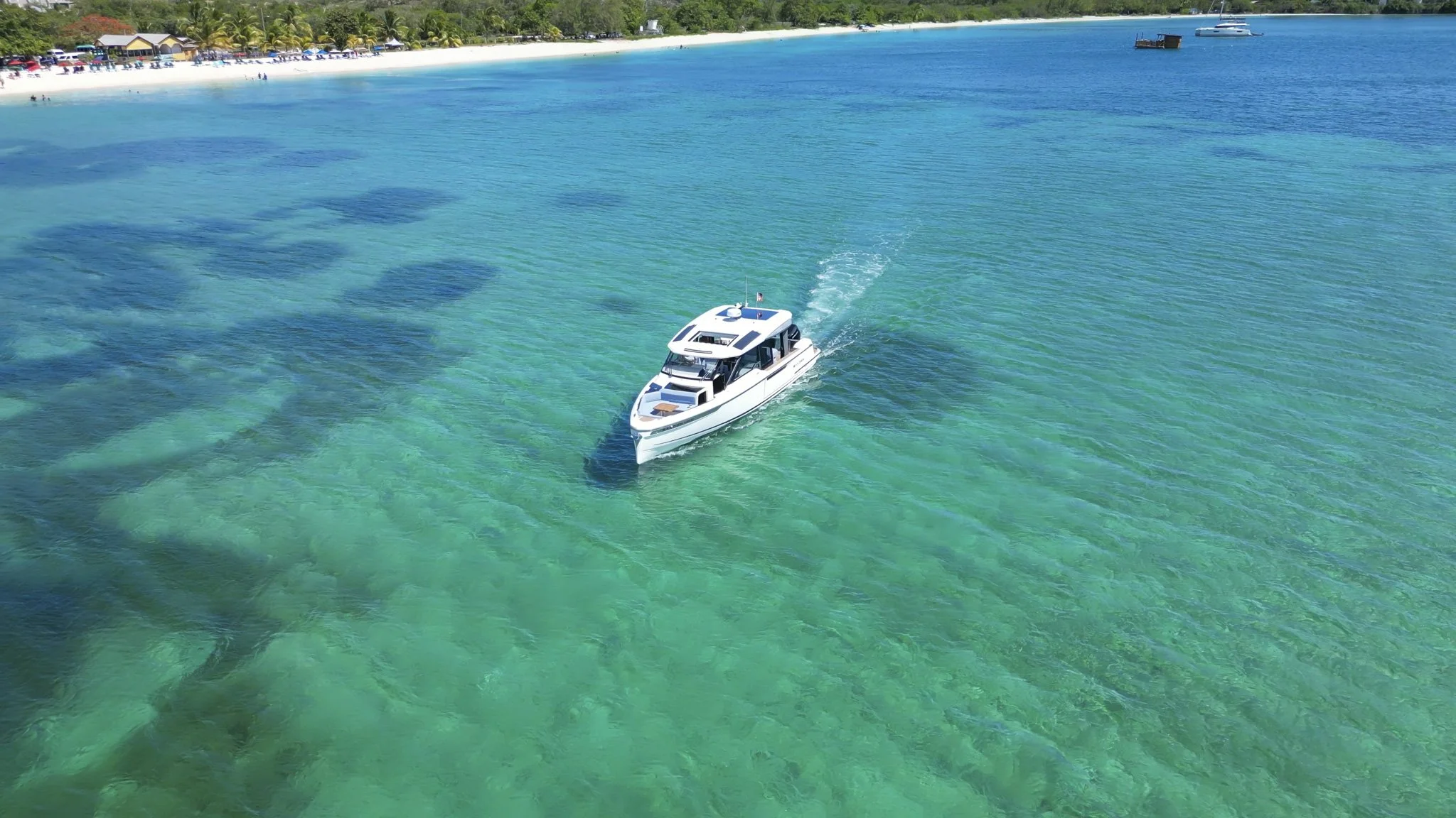 A white yacht sailing on clear turquoise waters near a tropical beach with people and umbrellas, and lush green trees in the background.