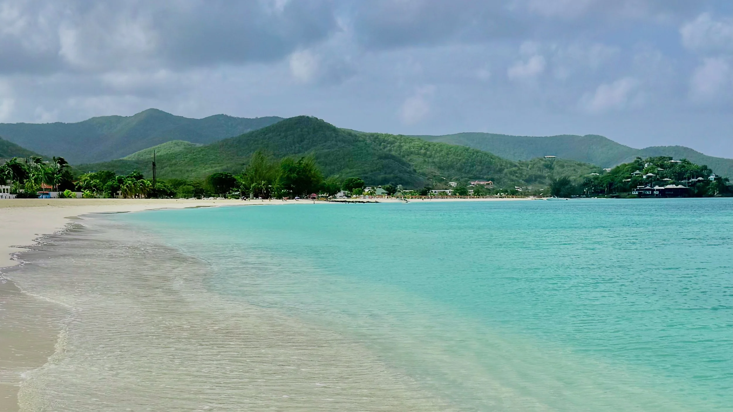 A tropical beach with white sand, clear turquoise water, lush green hills in the background, and a cloudy sky over the scene.