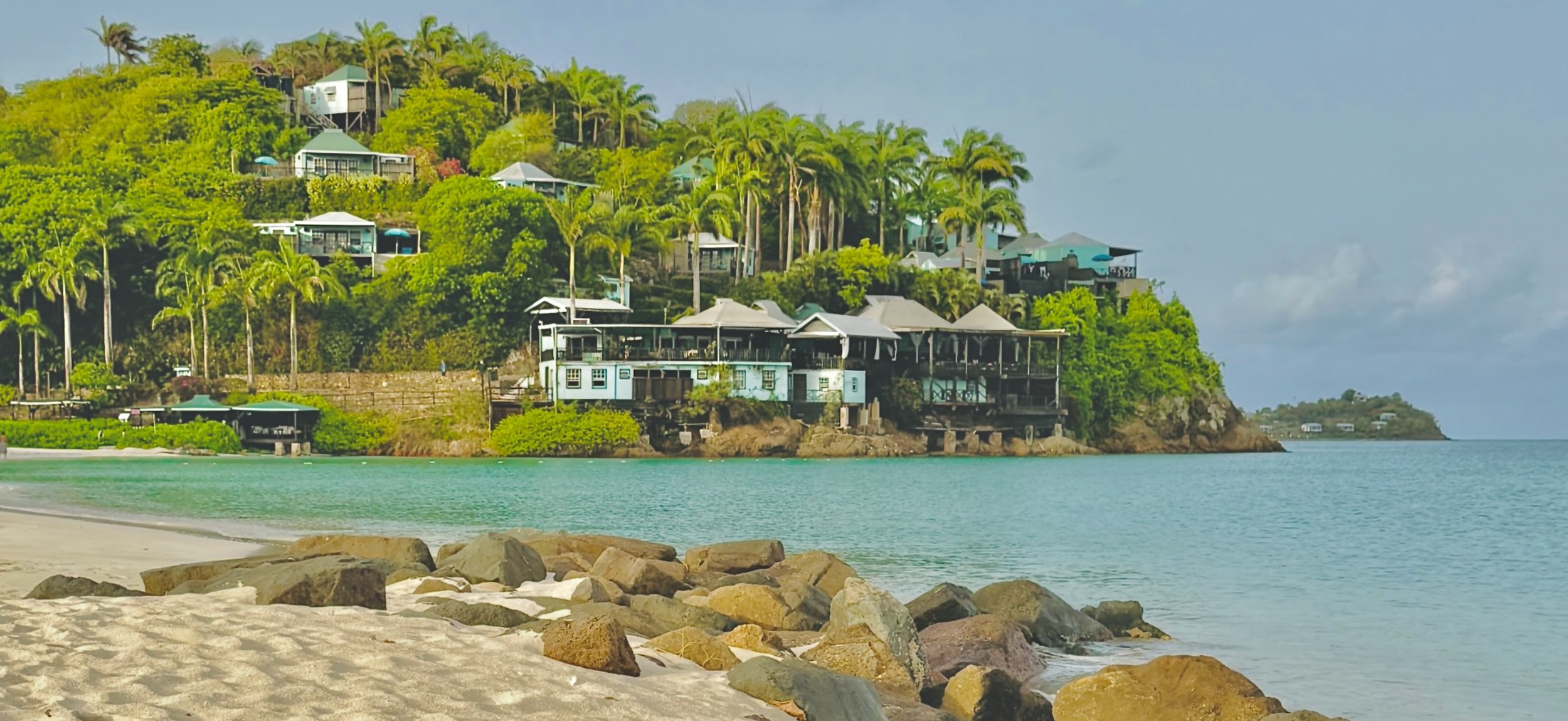 A tropical hillside with multiple houses and villas overlooking a calm, turquoise bay, with a sandy beach and rocks in the foreground.